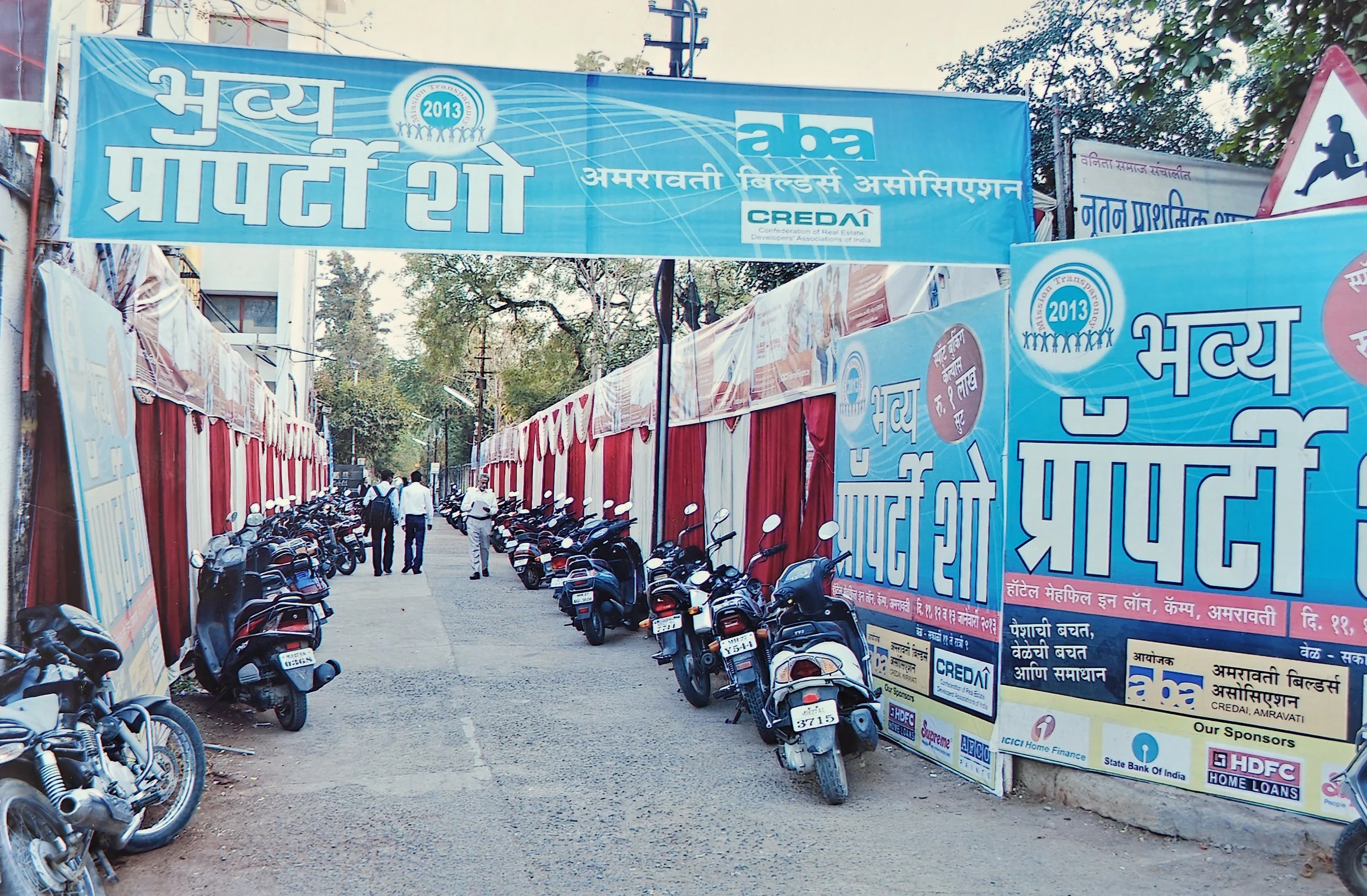 An outdoor street scene with parked motorcycles on both sides of a narrow dirt road, red and white fabric barriers, and large blue banners with Hindi text and logos, including '2013', 'aba', and 'CREDAI'.