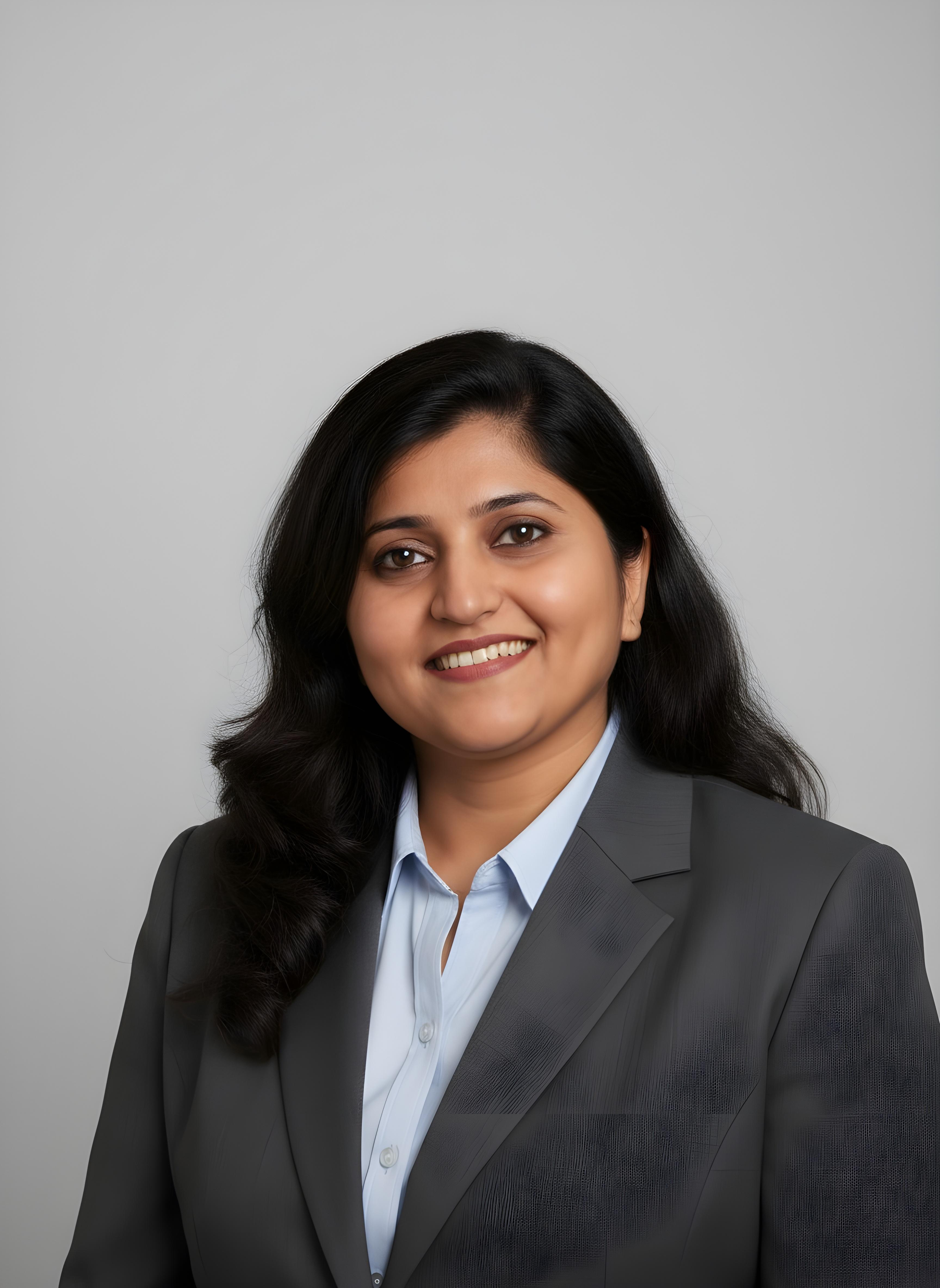 Professional portrait of a woman with long black hair wearing a gray blazer and light blue shirt, smiling on a plain light gray background.
