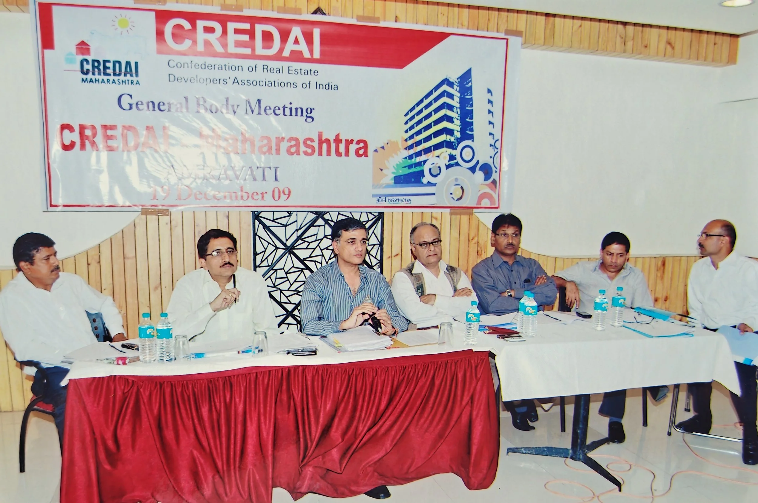 A group of seven men sitting at a long table during a conference with a large banner that reads 'CREDAl Maharashtra' and details about a general body meeting. The men have bottled water, papers, and microphones in front of them.