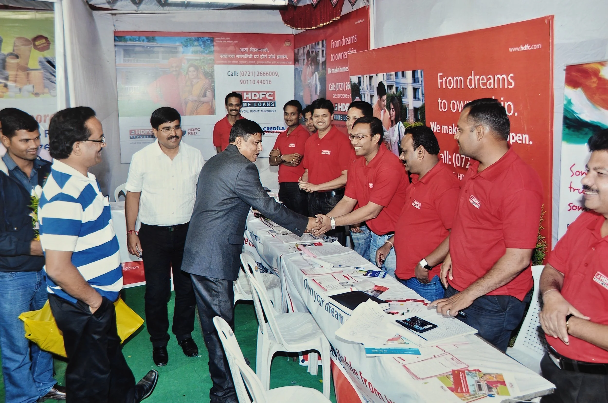 A group of people at a booth, some in red shirts, greeting a man in a suit with a handshake. The booth has banners and promotional materials related to HDFC and home loans.