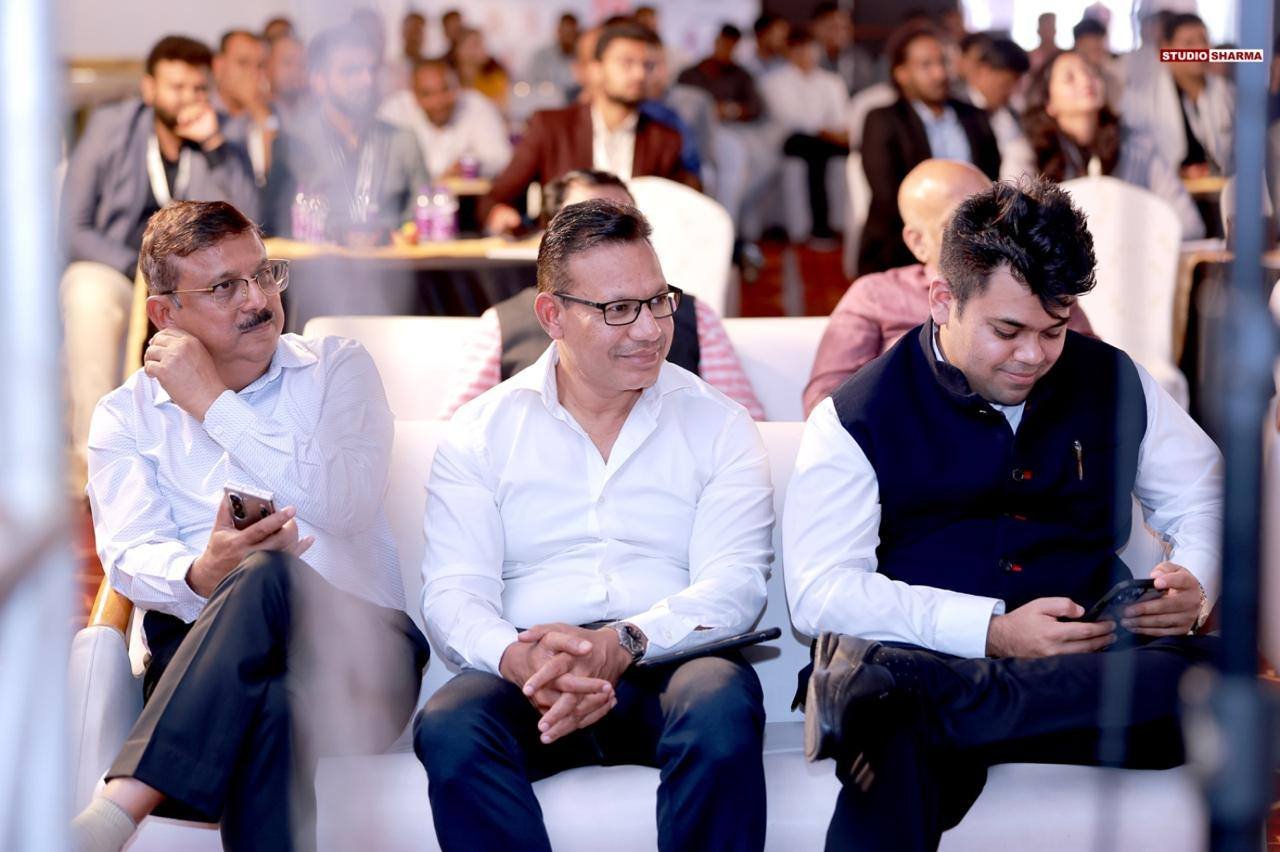 A group of people attending a conference or seminar, sitting on white sofas and chairs, some using their phones, with a large audience in the background.