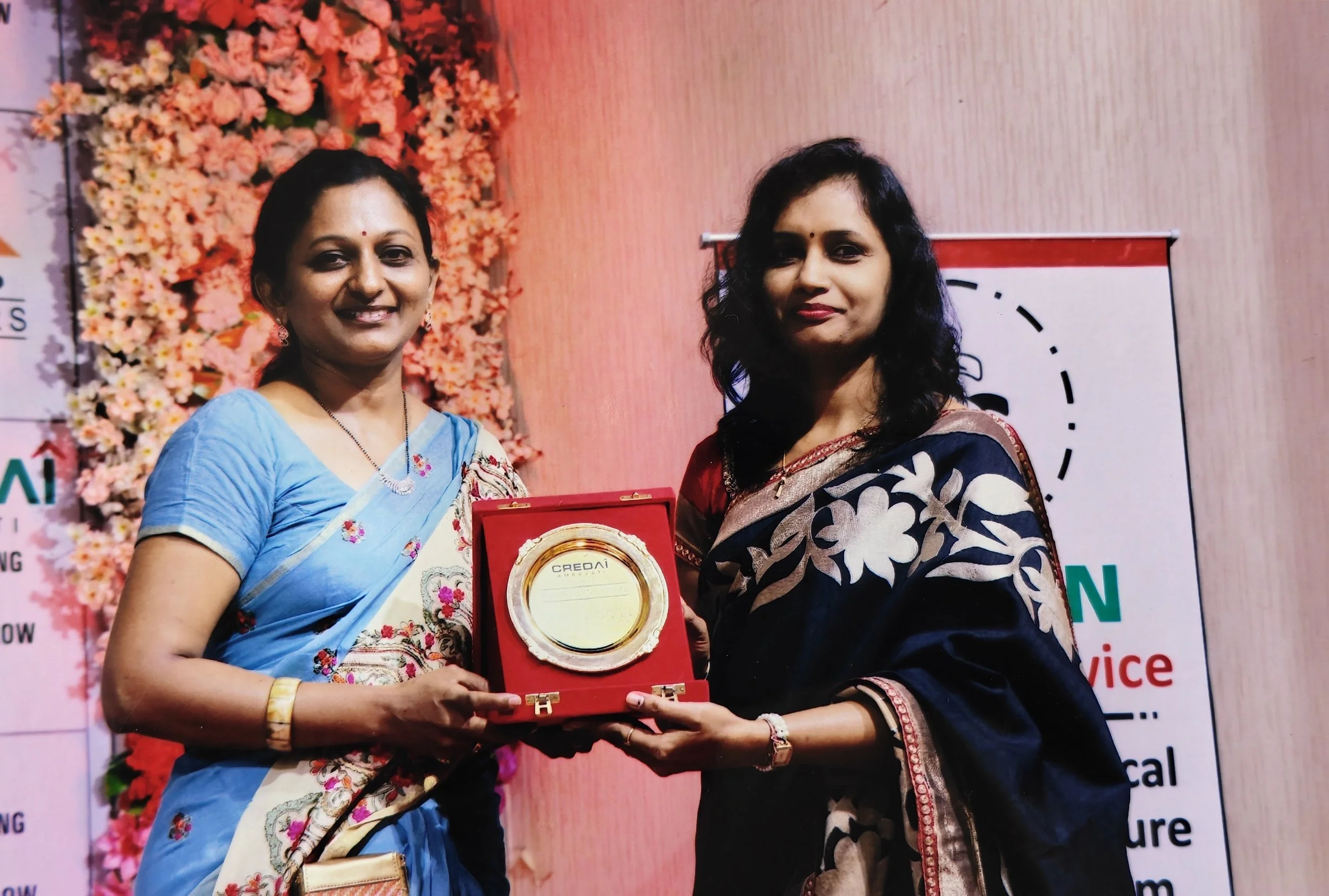 Two women in sarees standing together at an award ceremony, holding a plaque or award in a red box, with pink floral decorations and a banner in the background.