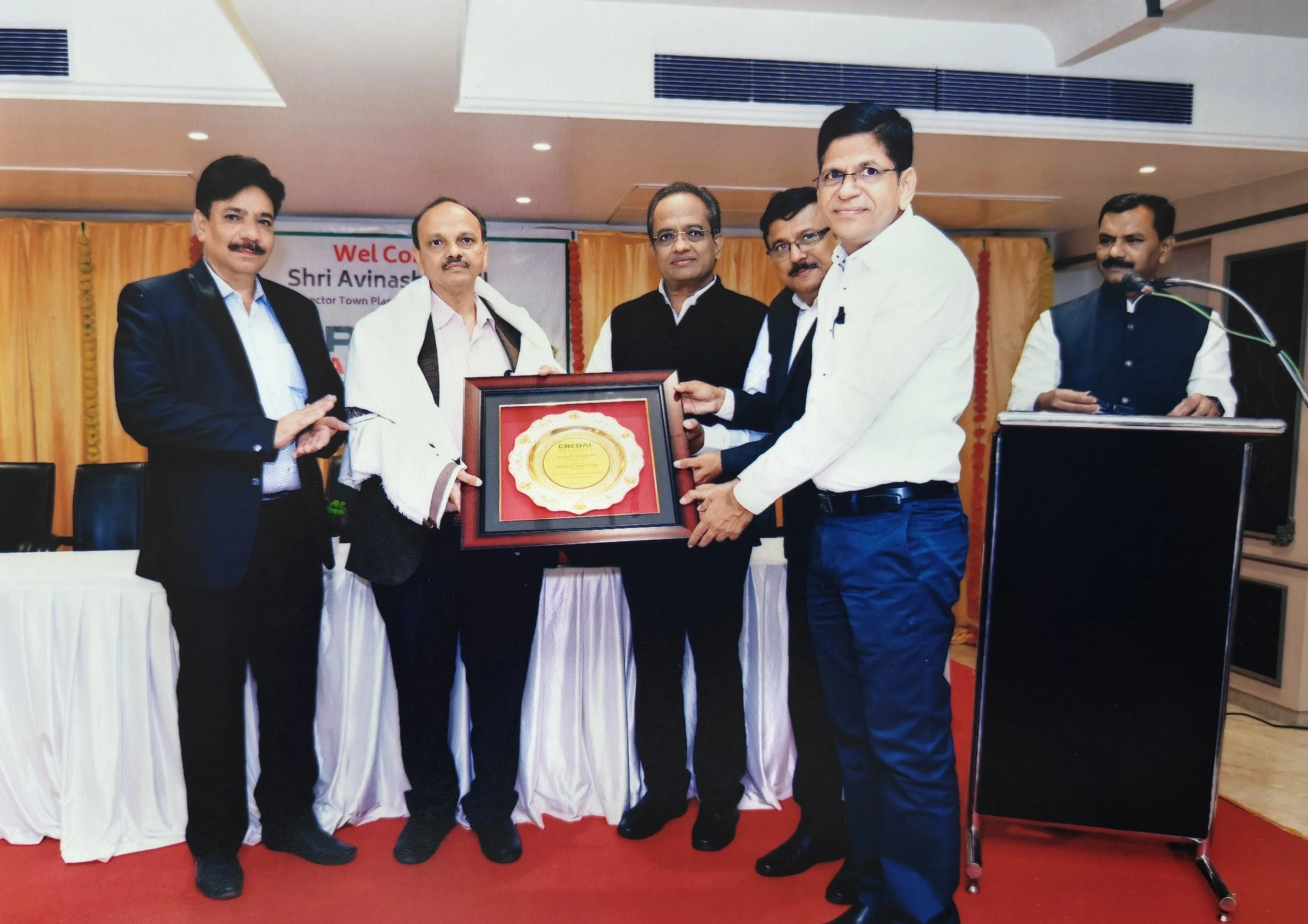 Five men in formal attire at an award ceremony, with one man presenting a framed ceremonial plaque to another, in front of a yellow curtain, some sitting tables, and a podium with a microphone.