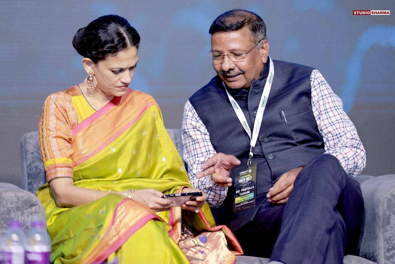 A woman in a yellow and pink saree and a man in a checkered shirt and vest sitting on a grey sofa, looking at a smartphone together at an event with a stage background. The woman appears focused on the phone while the man is talking and pointing at i