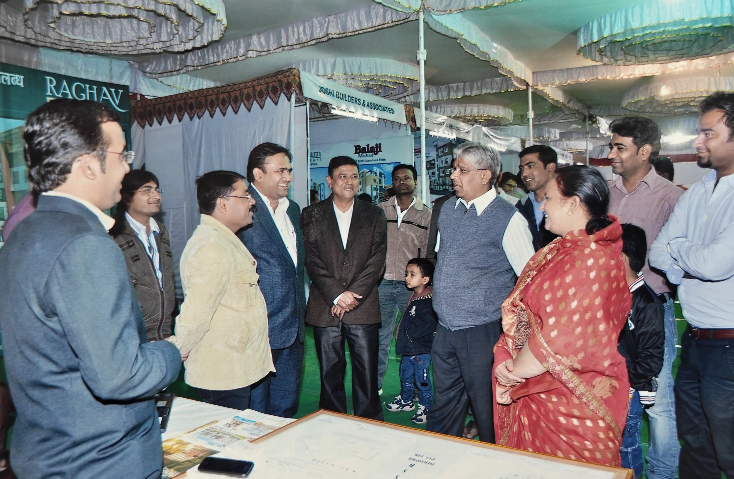 Group of people, including men, women, and children, gathered and talking under a decorated tent at an indoor event or exhibition. Most individuals are smiling and engaged in conversation.
