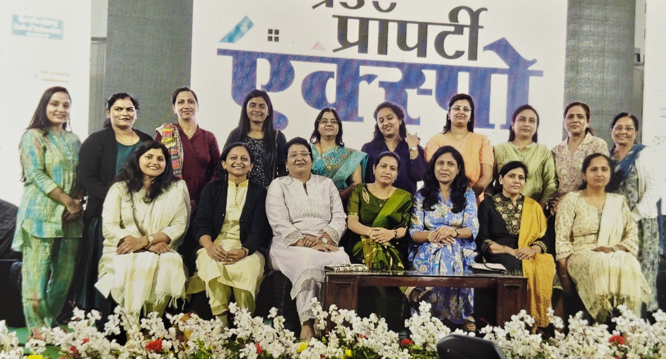 Group of women in traditional and semi-formal attire posing for a photo on stage with a large decorated backdrop in Hindi and floral decorations at the front.