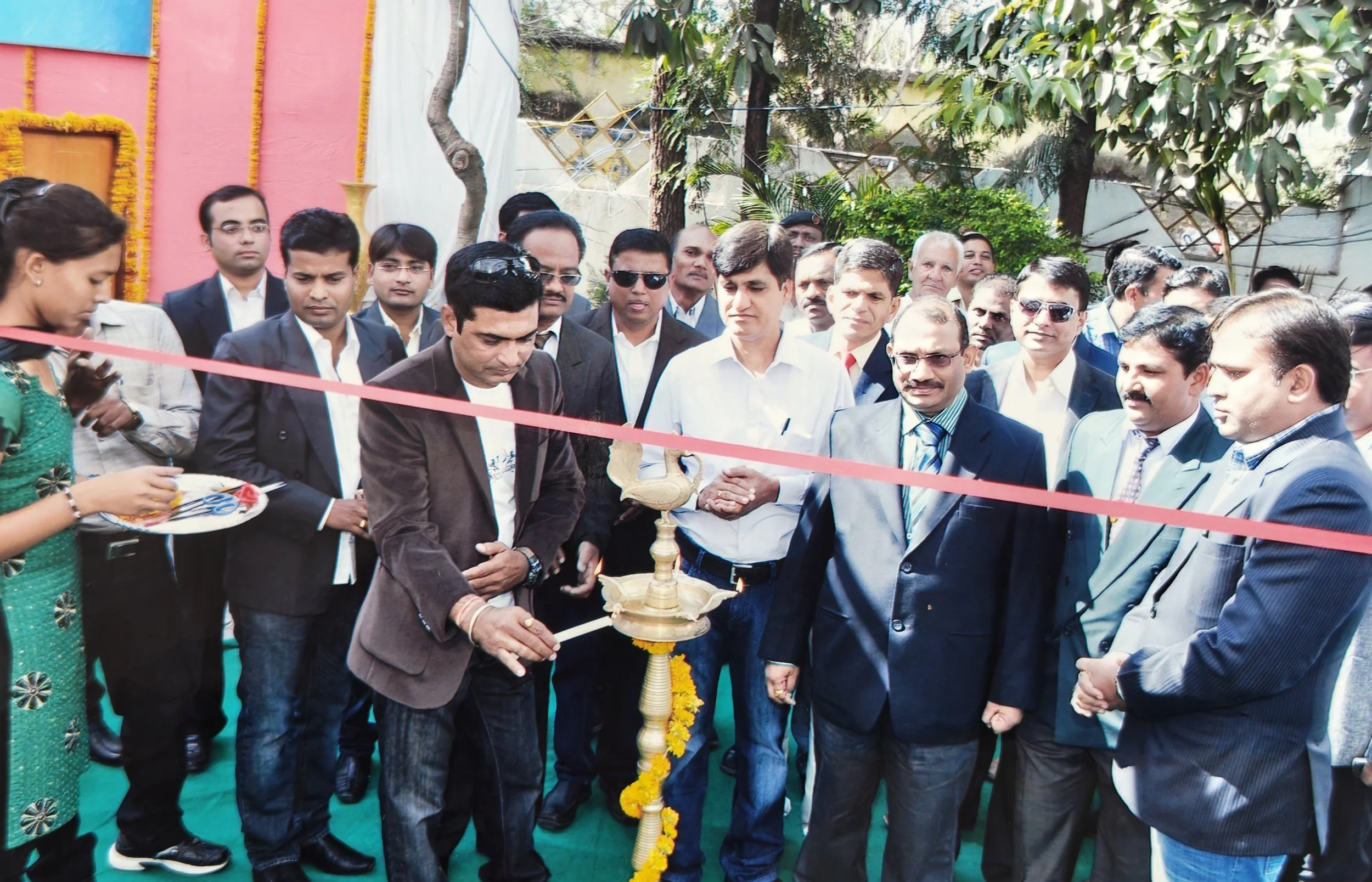 A group of men and women at an outdoor event, gathered around a traditional Indian lamp (diya) for a lamp-lighting ceremony, with a woman holding a tray with items on the left, and a red ribbon stretched across the scene.