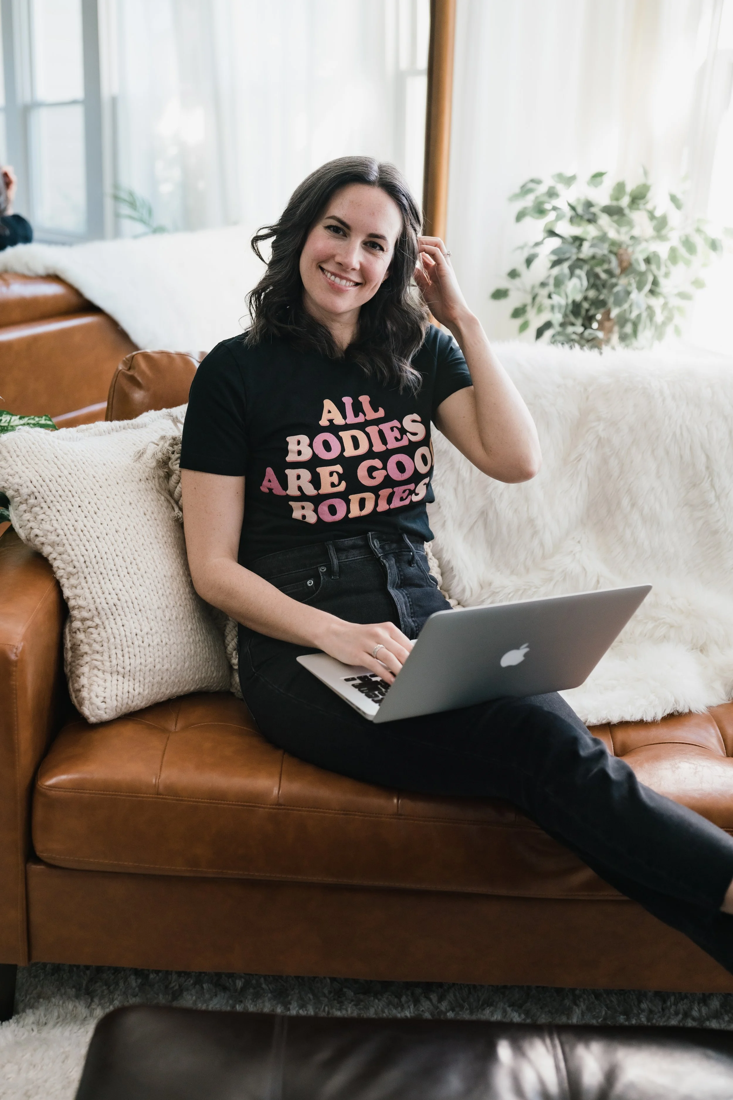 Kate Sutton smiling holding her hair back & sitting ona leather couch on a soft blanket. Kate Sutton offers eating disorder therapy & coaching for women wanting to heal their relationship with their body.