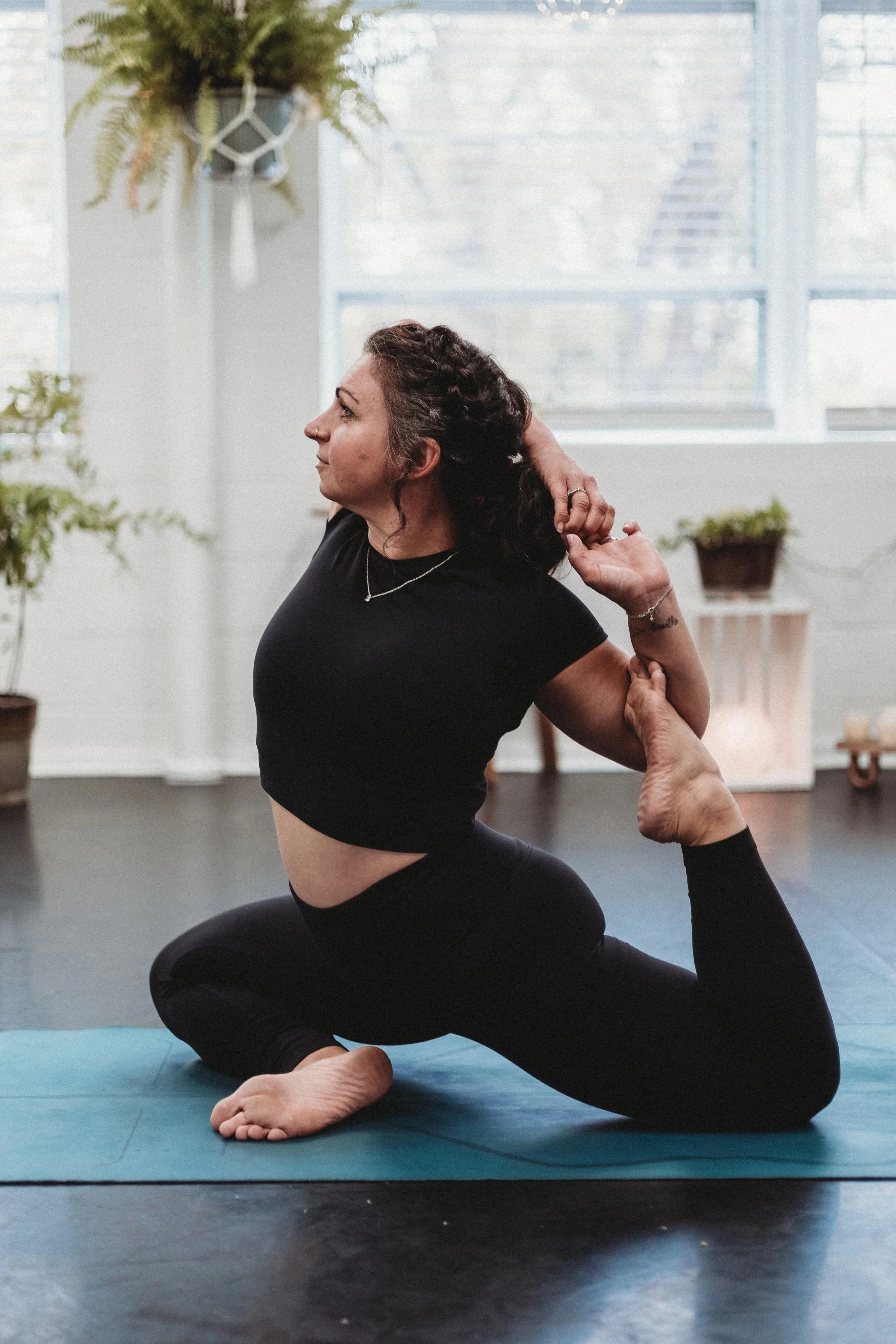 Woman practicing yoga indoors on a blue yoga mat, performing a backbend stretch, with potted plants and a large window in the background.