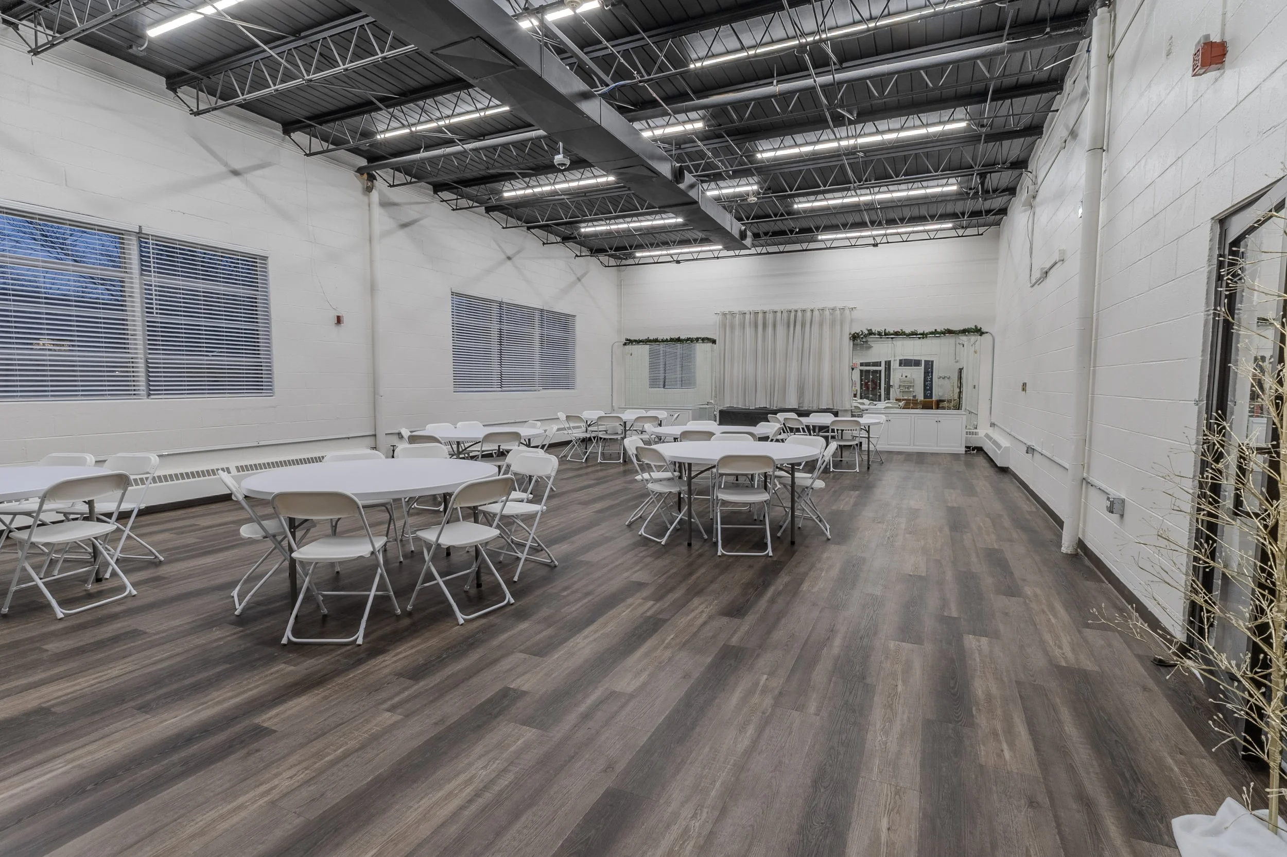 Event space with round tables and white folding chairs, a backdrop with curtains and string lights, and large windows with blinds.