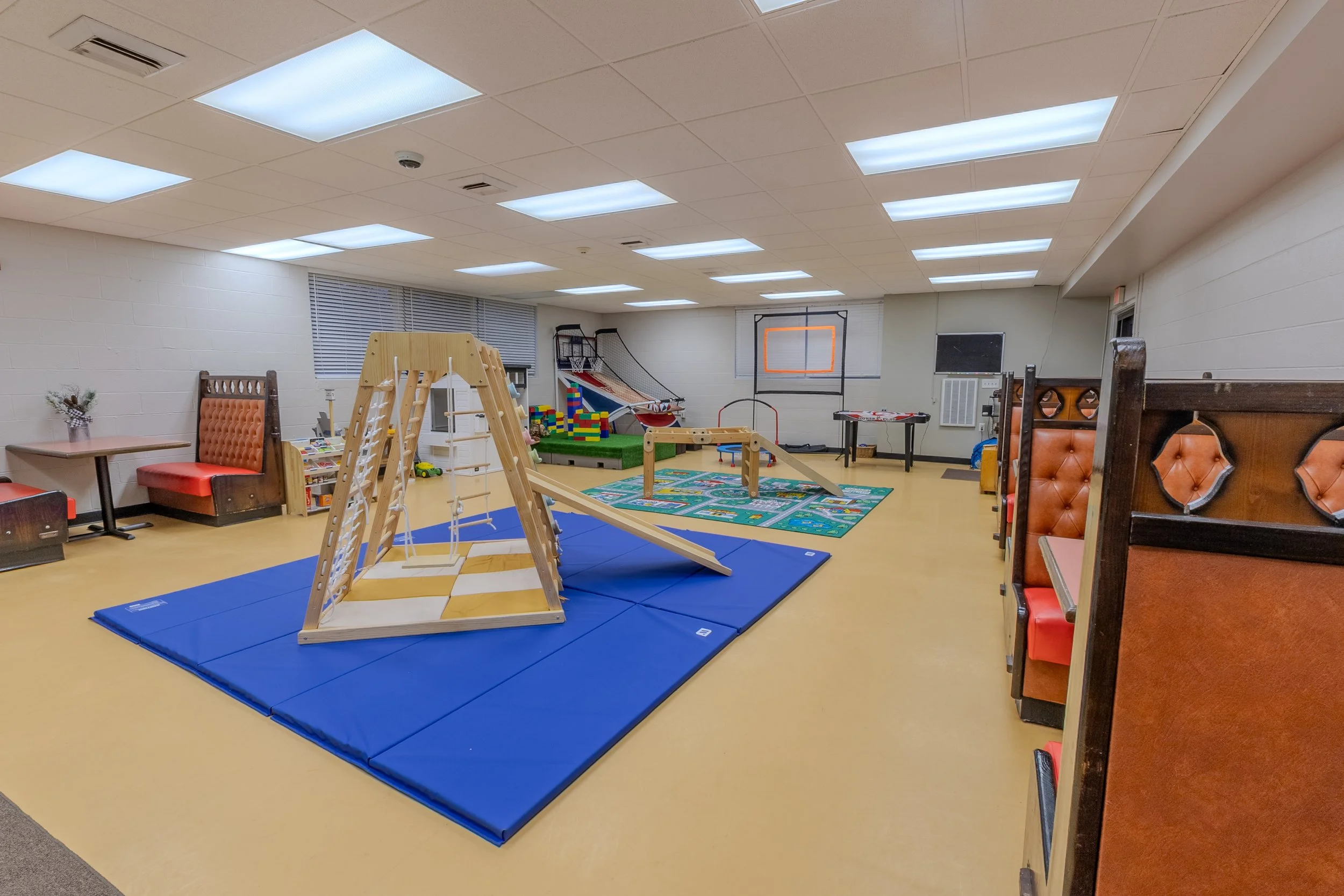 Children’s play area with colorful rug, small slide, tables with booths, arcade game, basketball hoop, and other toys.