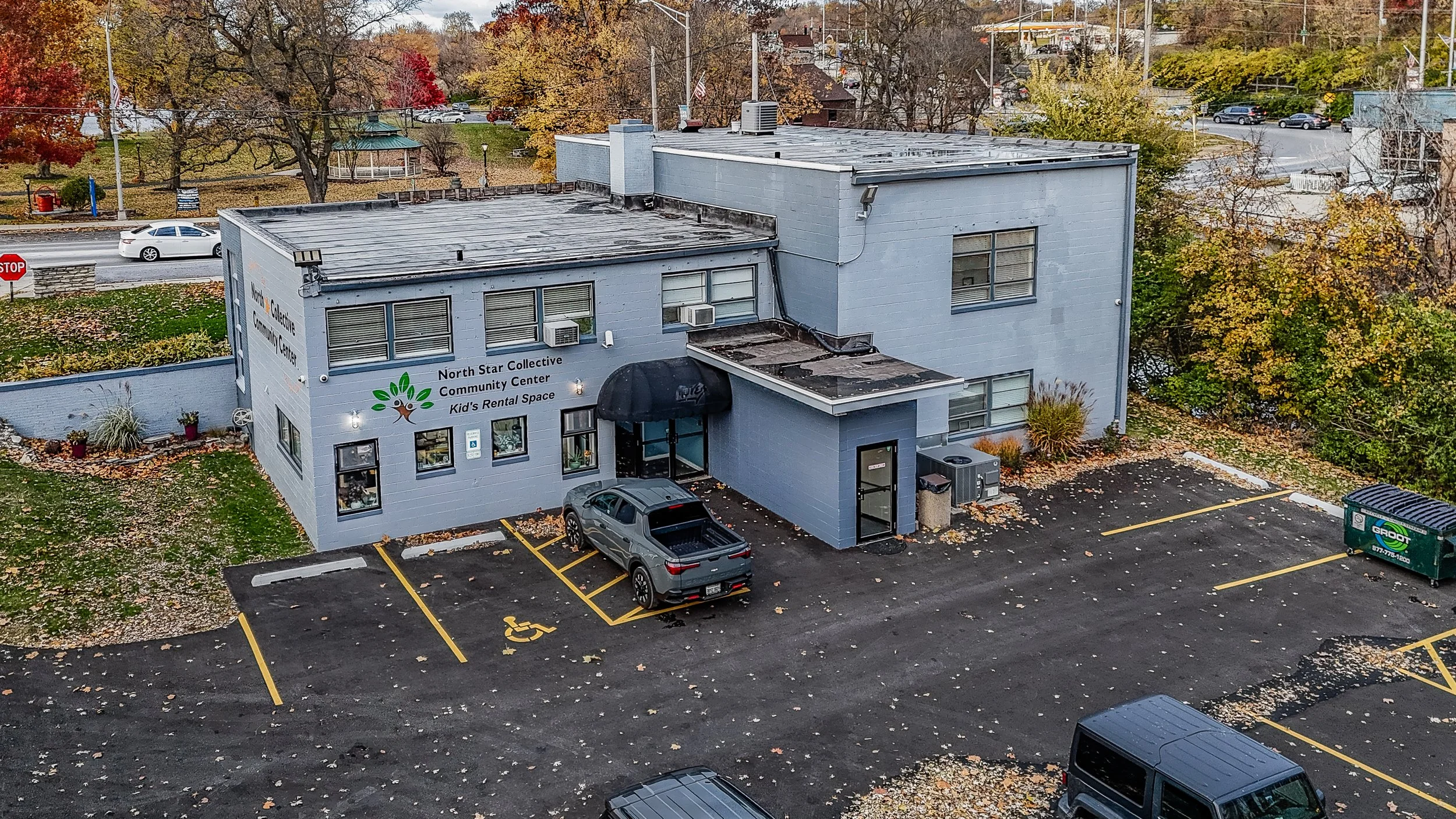 An aerial view of a two-story light blue community center building with parking lot, including a reserved spot for handicapped parking. The building has a sign that reads "North Star Collective Community Center, Kid's Rental Space" and features a logo with a tree and green leaves. Surrounding trees have autumn leaves, and traffic with cars is visible on the street behind the building.