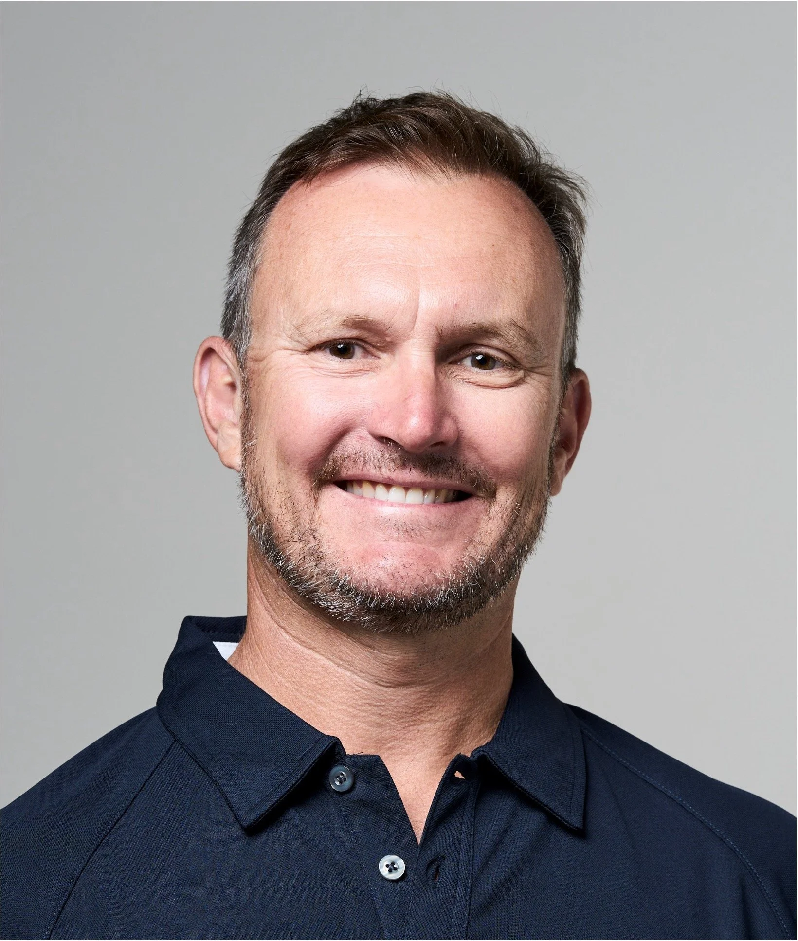 A middle-aged man with short hair and a beard, smiling, wearing a navy blue collared shirt, in front of a plain light gray background.
