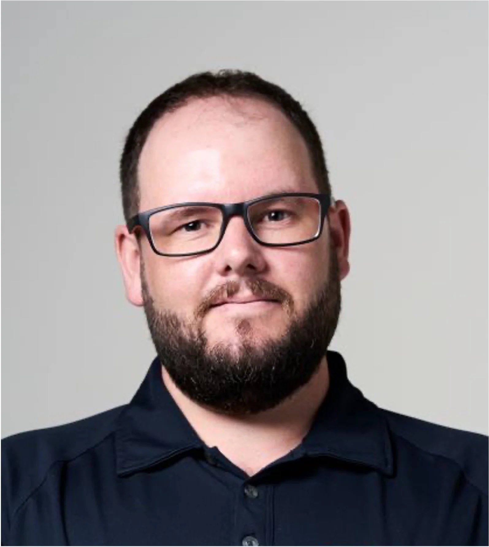 Portrait of a man with glasses and a beard wearing a dark collared shirt against a light background.