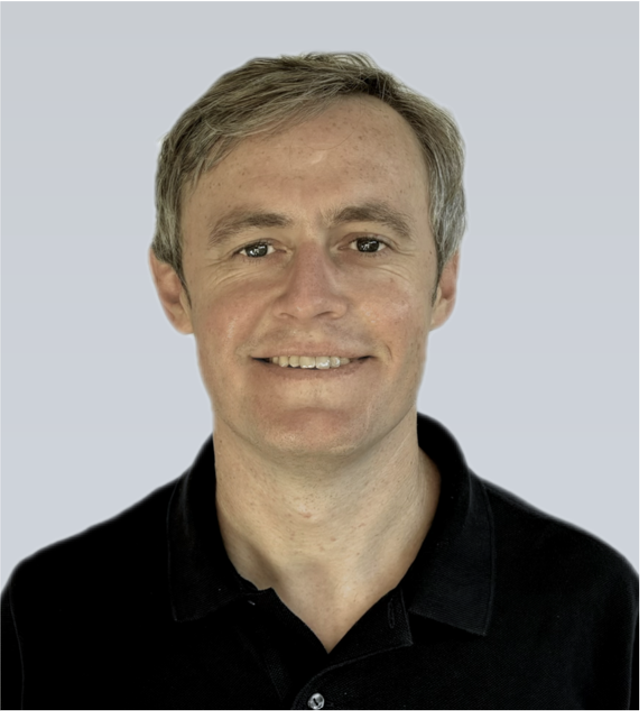 A young Caucasian man with short blonde hair smiling, wearing a black polo shirt, against a plain light-colored background.