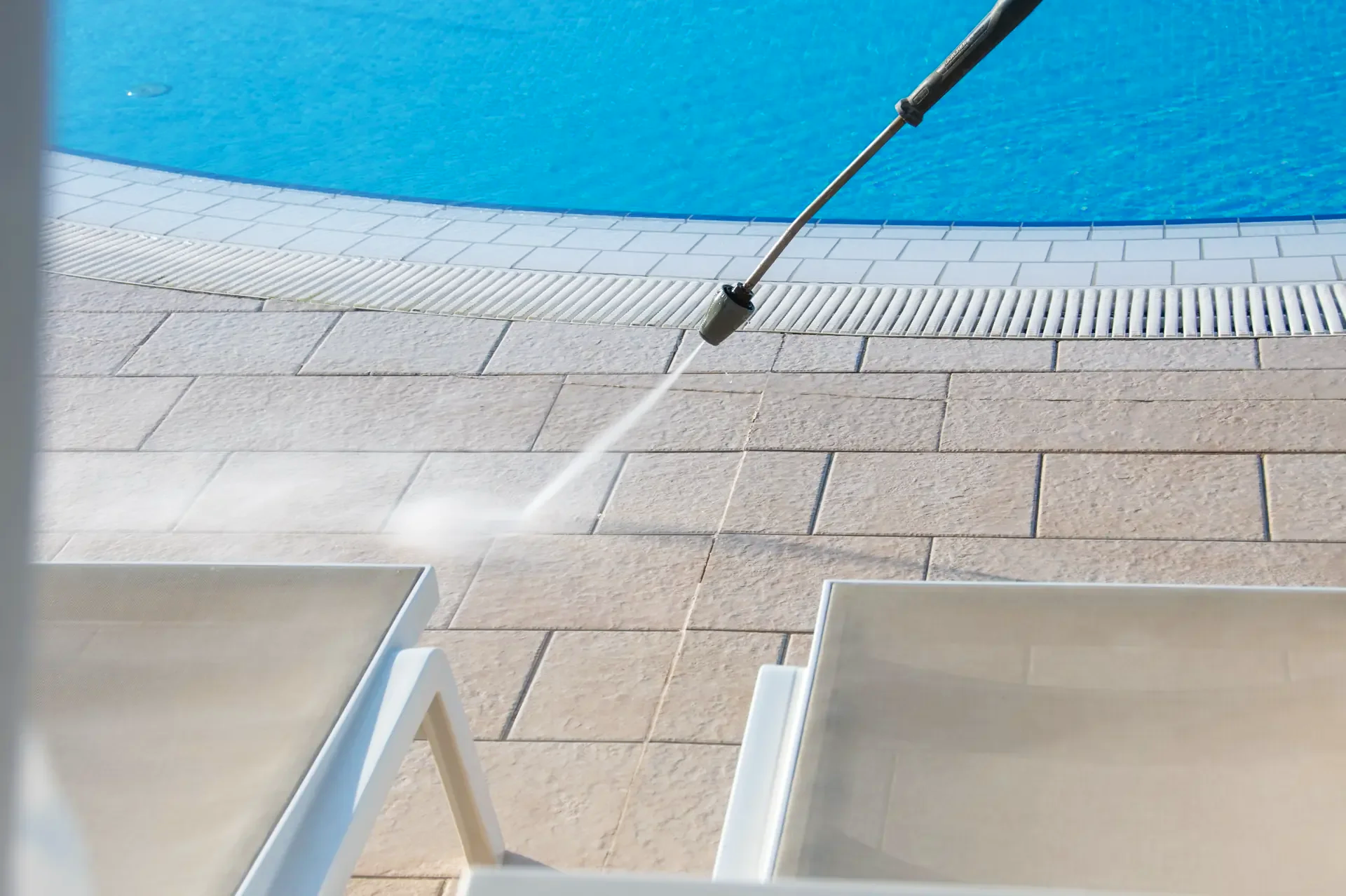 Poolside area with a spray gun cleaning beige tiled floor near a blue swimming pool, with two white lounge chairs in the foreground.
