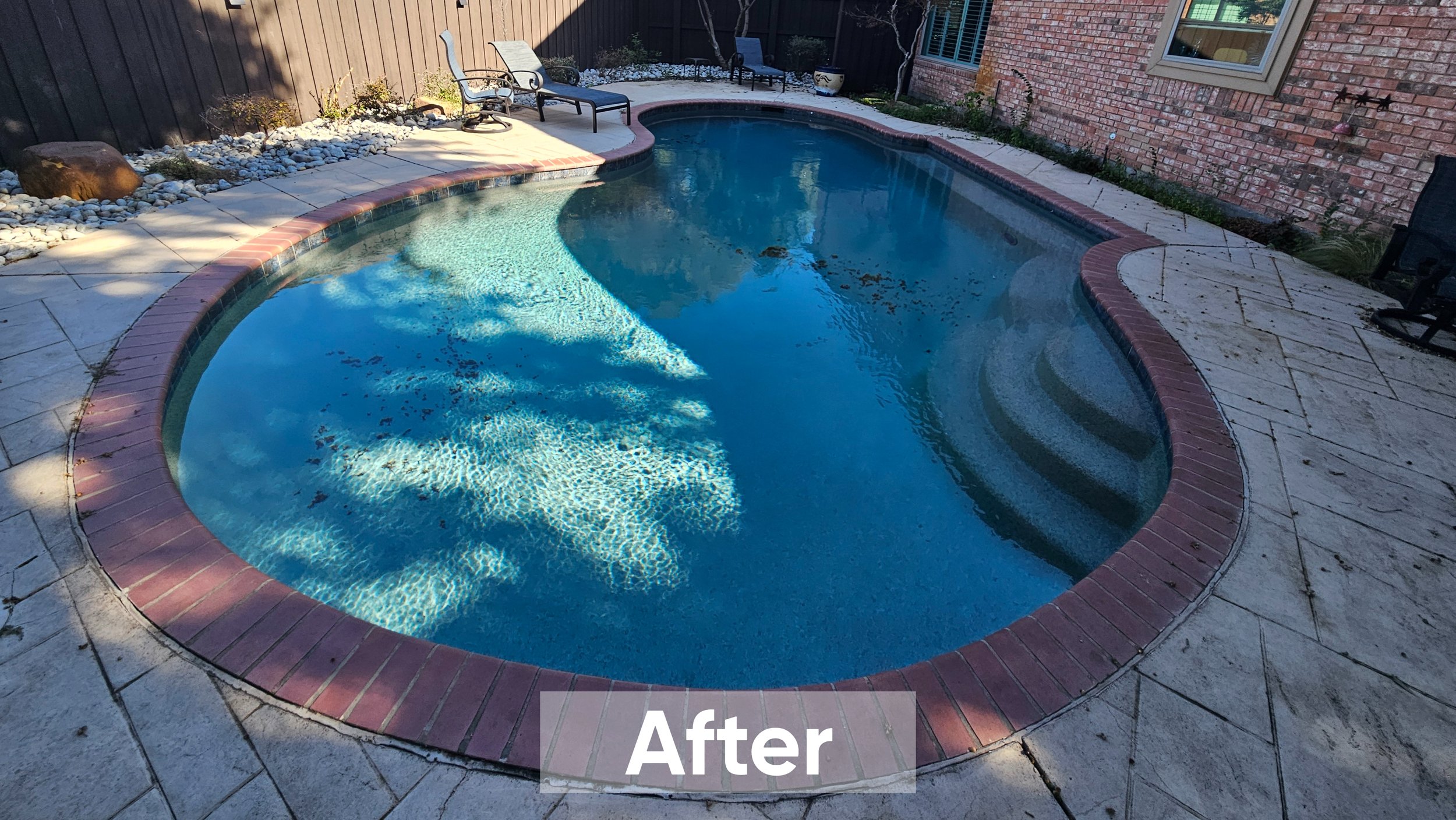 Clean, shaded kidney-shaped backyard pool with steps, surrounded by decorative bricks and light-colored stone paving, with patio chairs and a rock garden in the background.