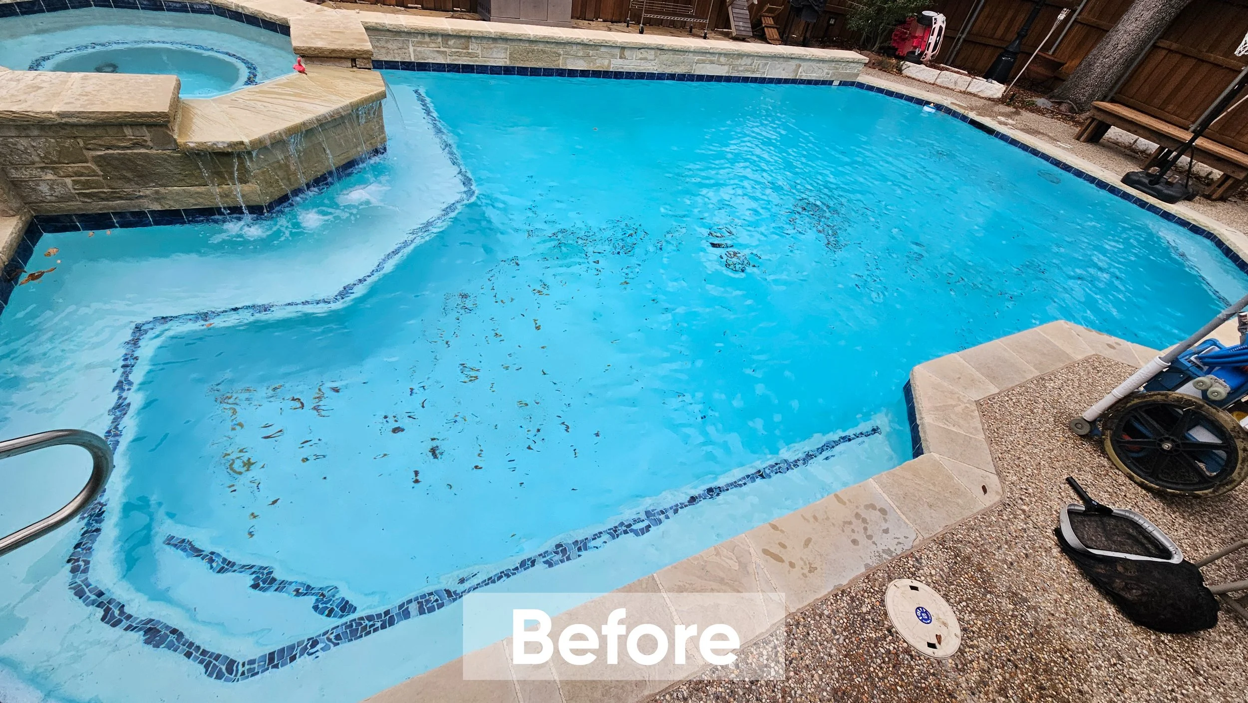 An outdoor swimming pool with a spa feature in a backyard, surrounded by a concrete deck and wooden fencing, with a pool cleaning net and a roller nearby.
