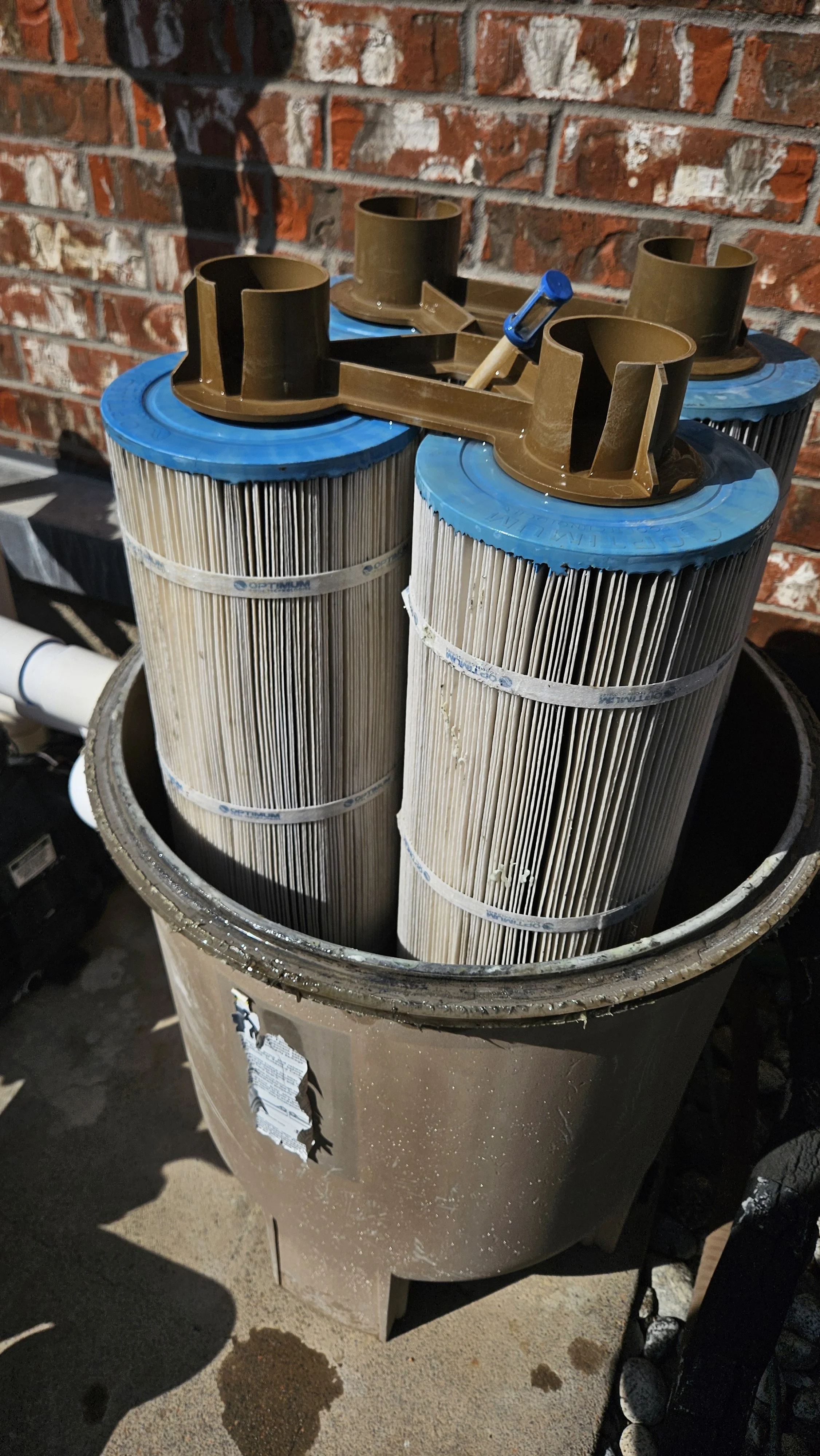 Pool filter cartridges in a brown trash bin outside against a brick wall, with a small blue-handled tool on top.