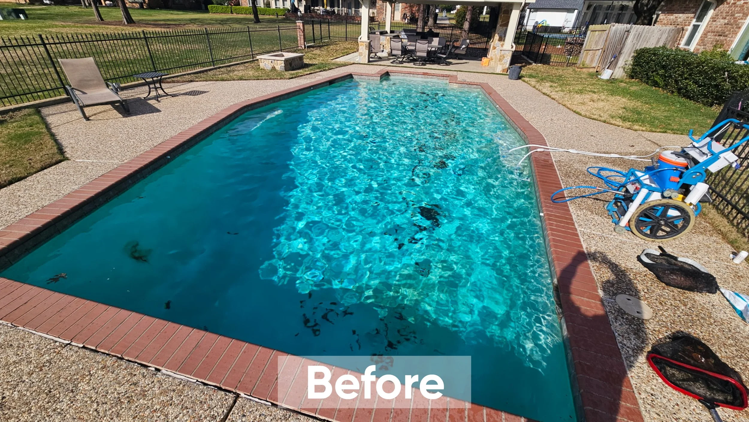 A backyard swimming pool with clear blue water, surrounded by a brick border and textured concrete decking. There are a few items around the pool, including a pool cleaning robot, a black bag, and some hoses. The backyard has a lawn, outdoor furnitur