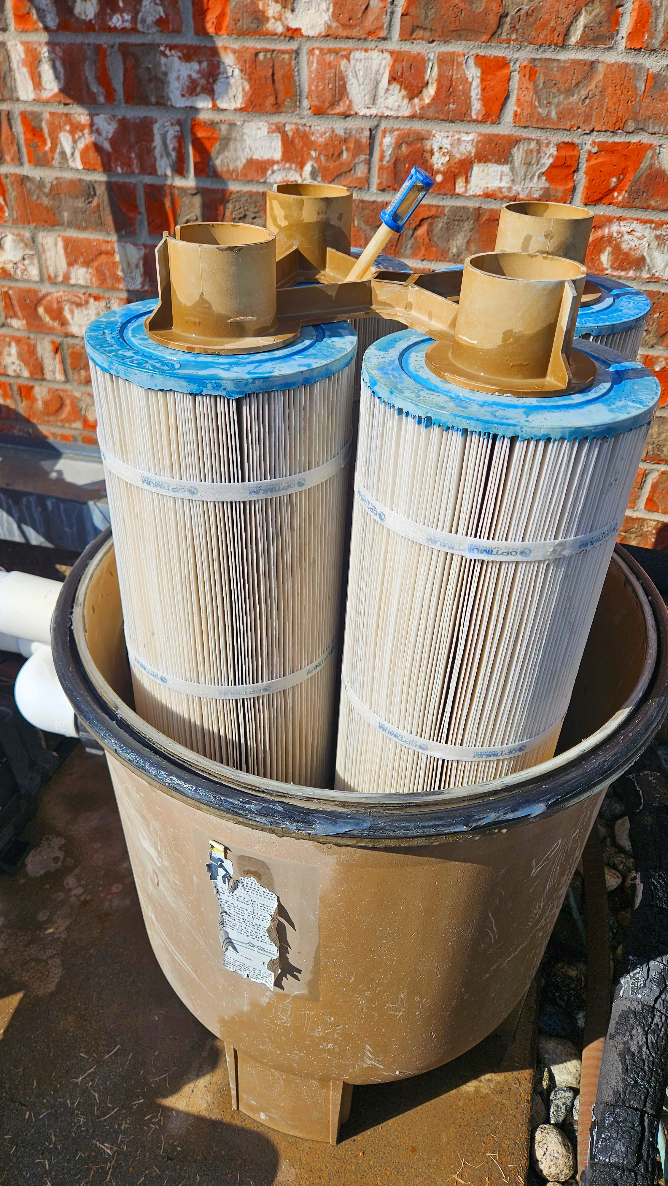 Large water filter clean cartridges stored in a beige plastic drum with a brick wall in the background.