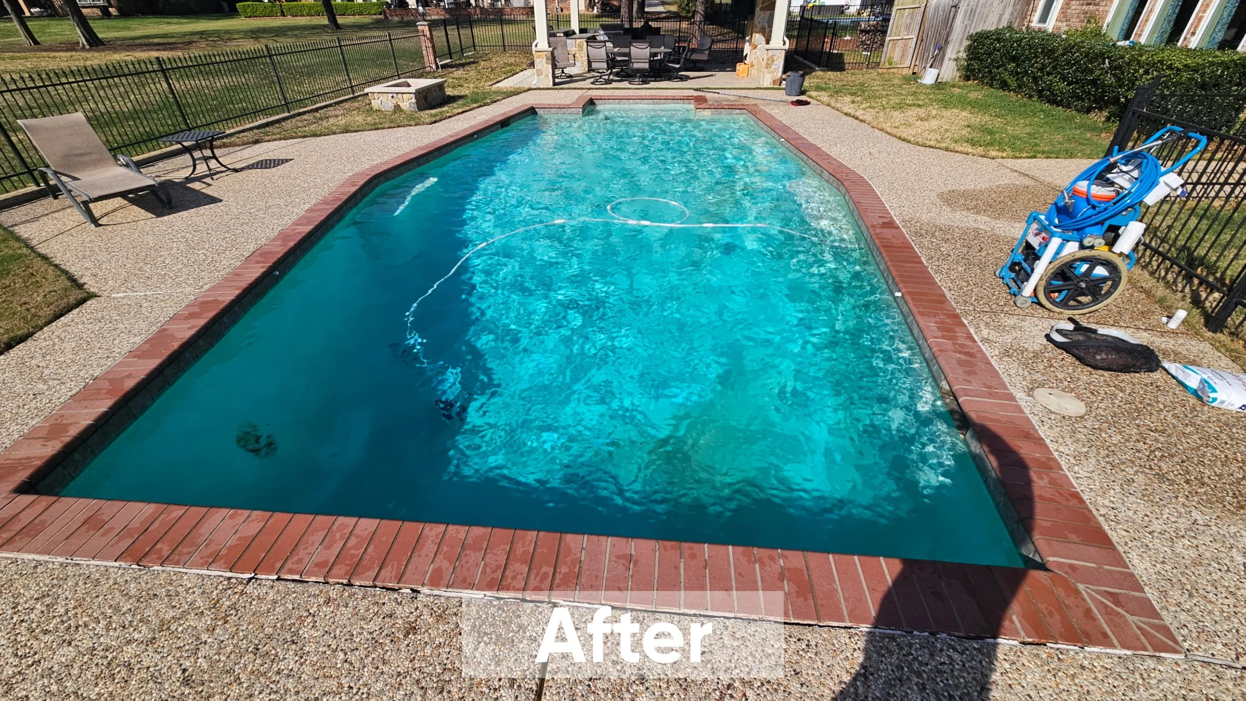 Clean in-ground swimming pool with clear blue water, surrounded by a brick border and concrete decking. Pool cleaning equipment and furniture are visible, with a fenced yard and patio furniture in the background.