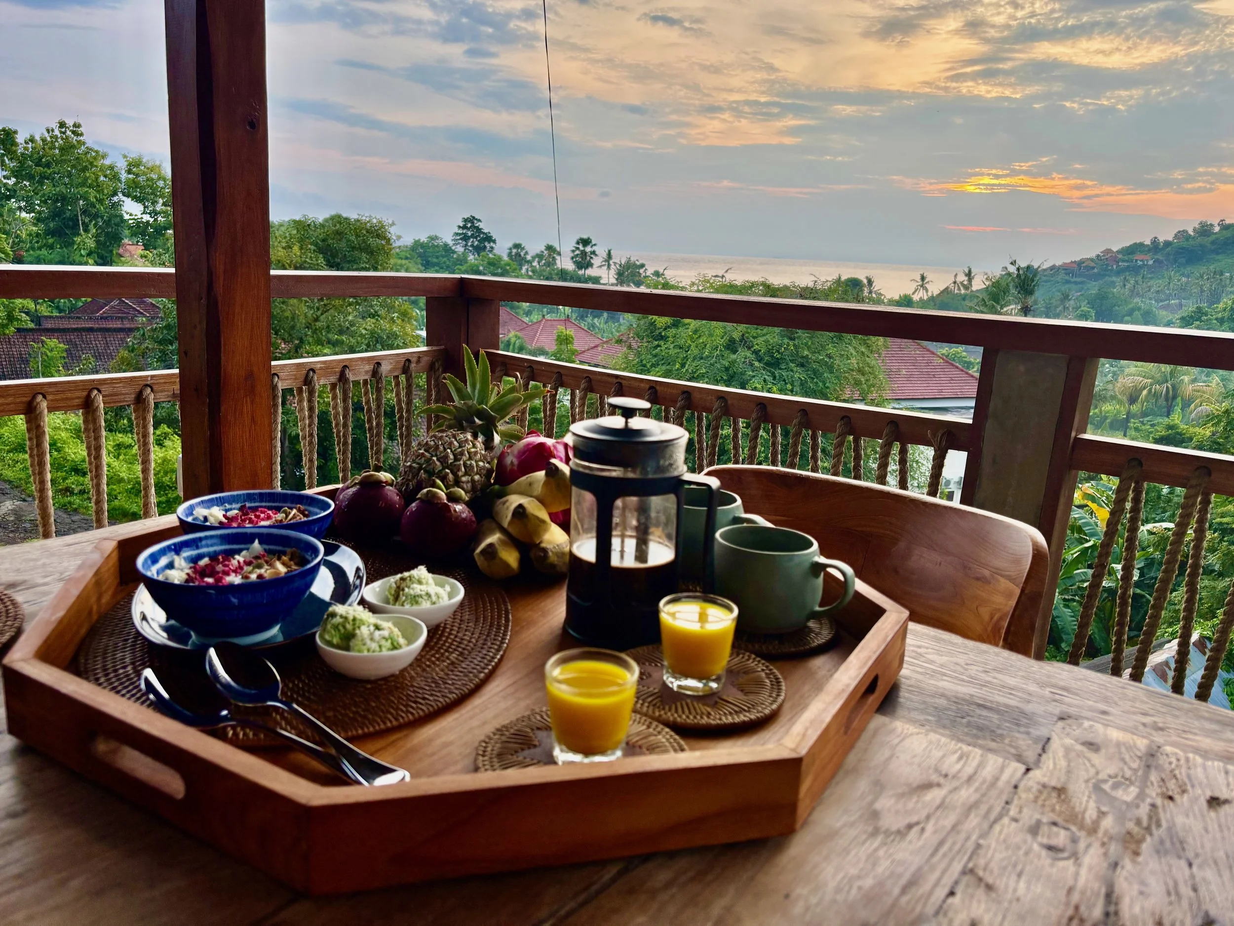Breakfast on a balcony table overlooking tropical landscape and sunset, with bowls of fruit, juice, coffee, and a French press.