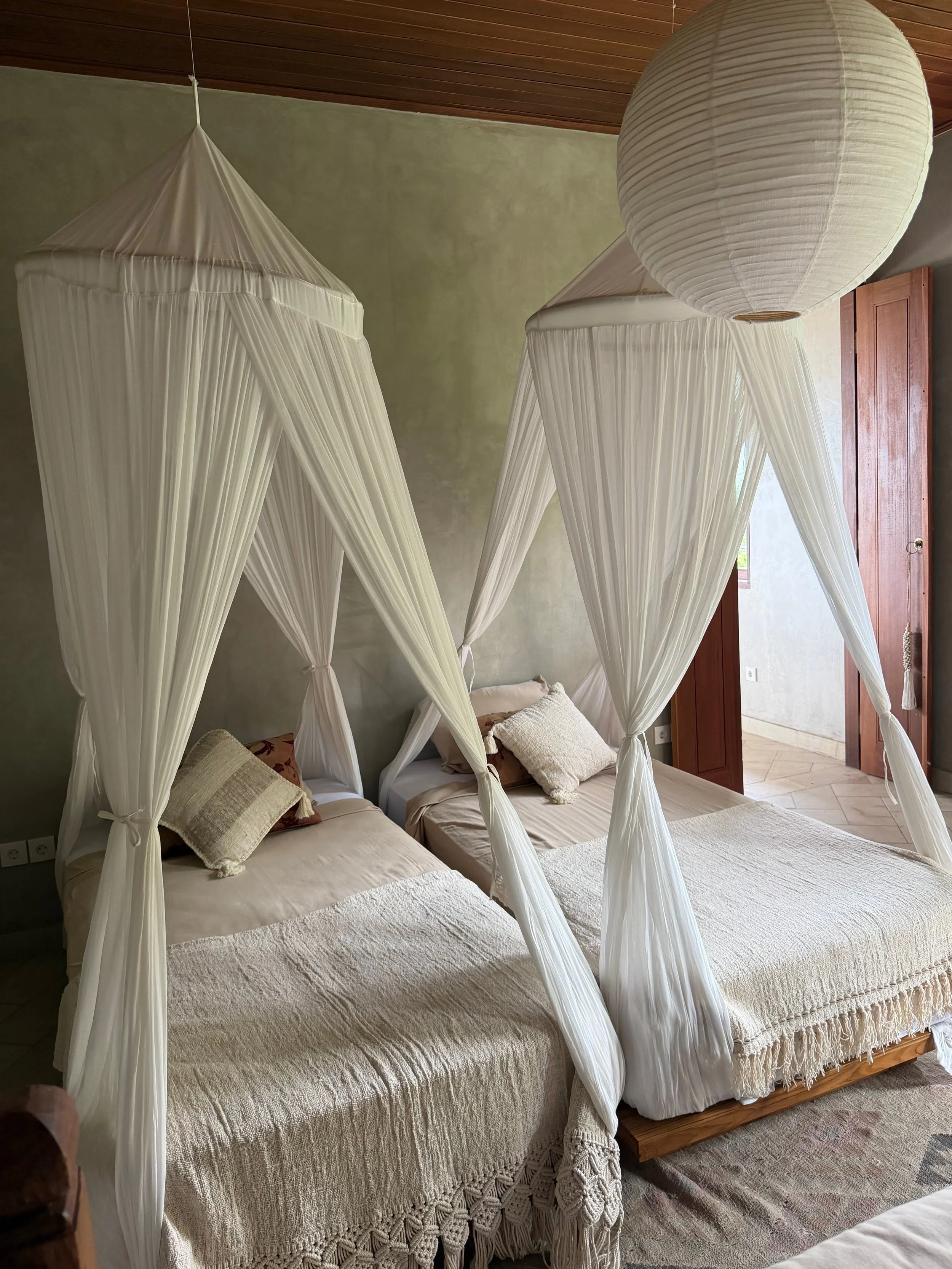 Two beds with white canopies in a room with wooden ceiling and open doorway.