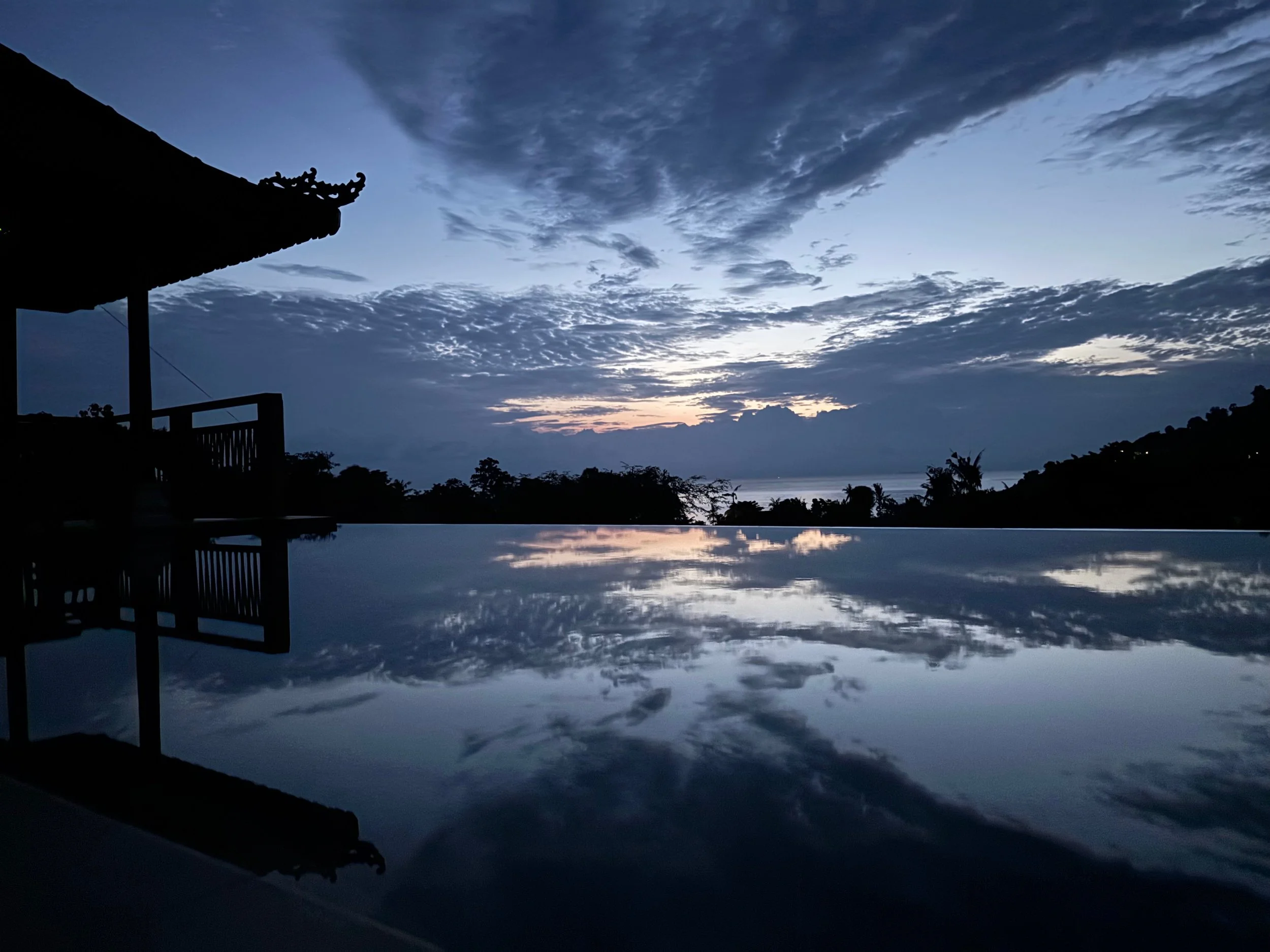 Sunset over a body of water with clouds in the sky, silhouette of a traditional Asian building on the left, and the water's surface reflecting the sky and clouds.