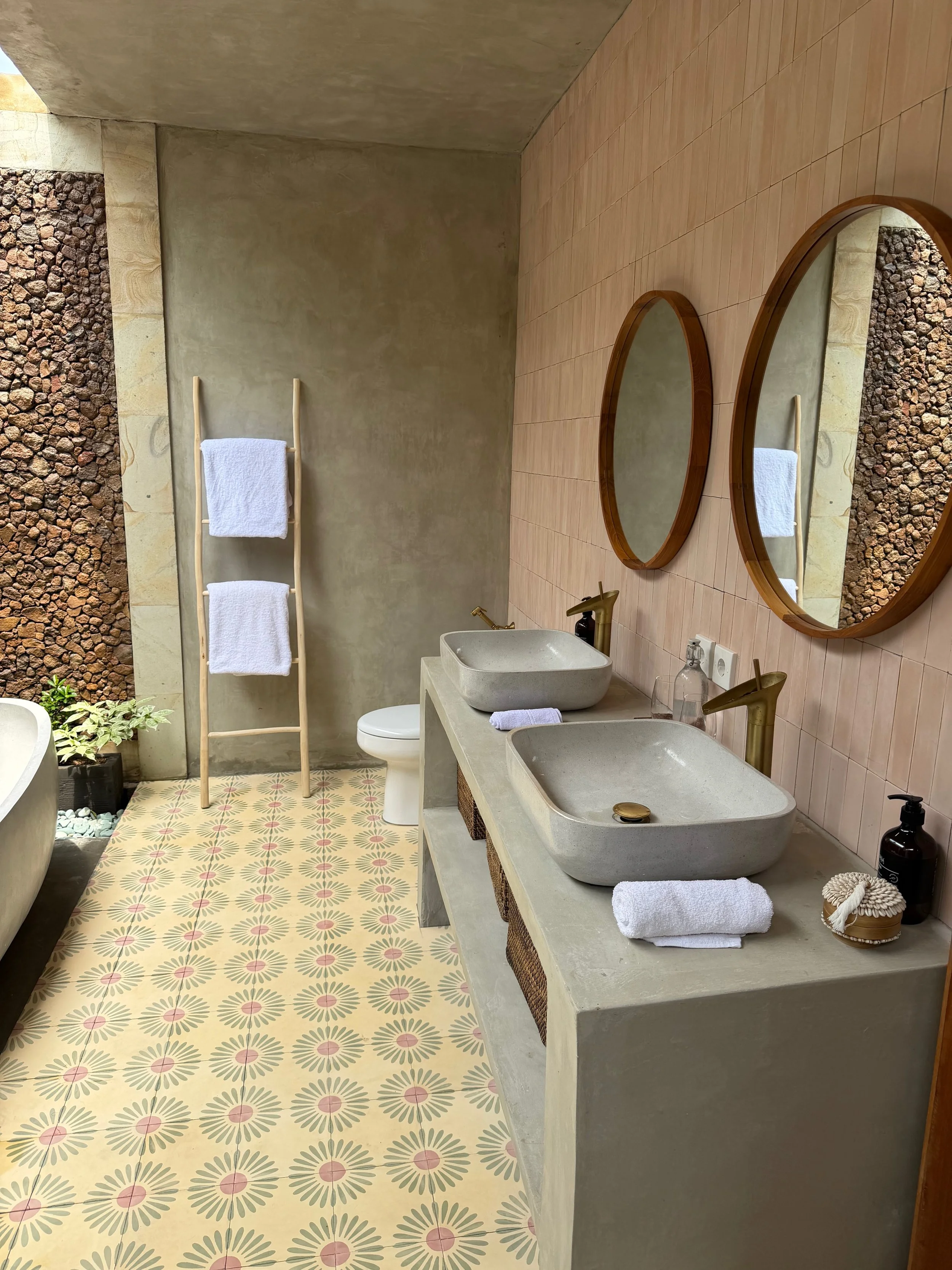 Bathroom with two vessel sinks, round mirrors, towel rack with white towels, bathtub, patterned tile floor, potted plant, and a concrete wall.