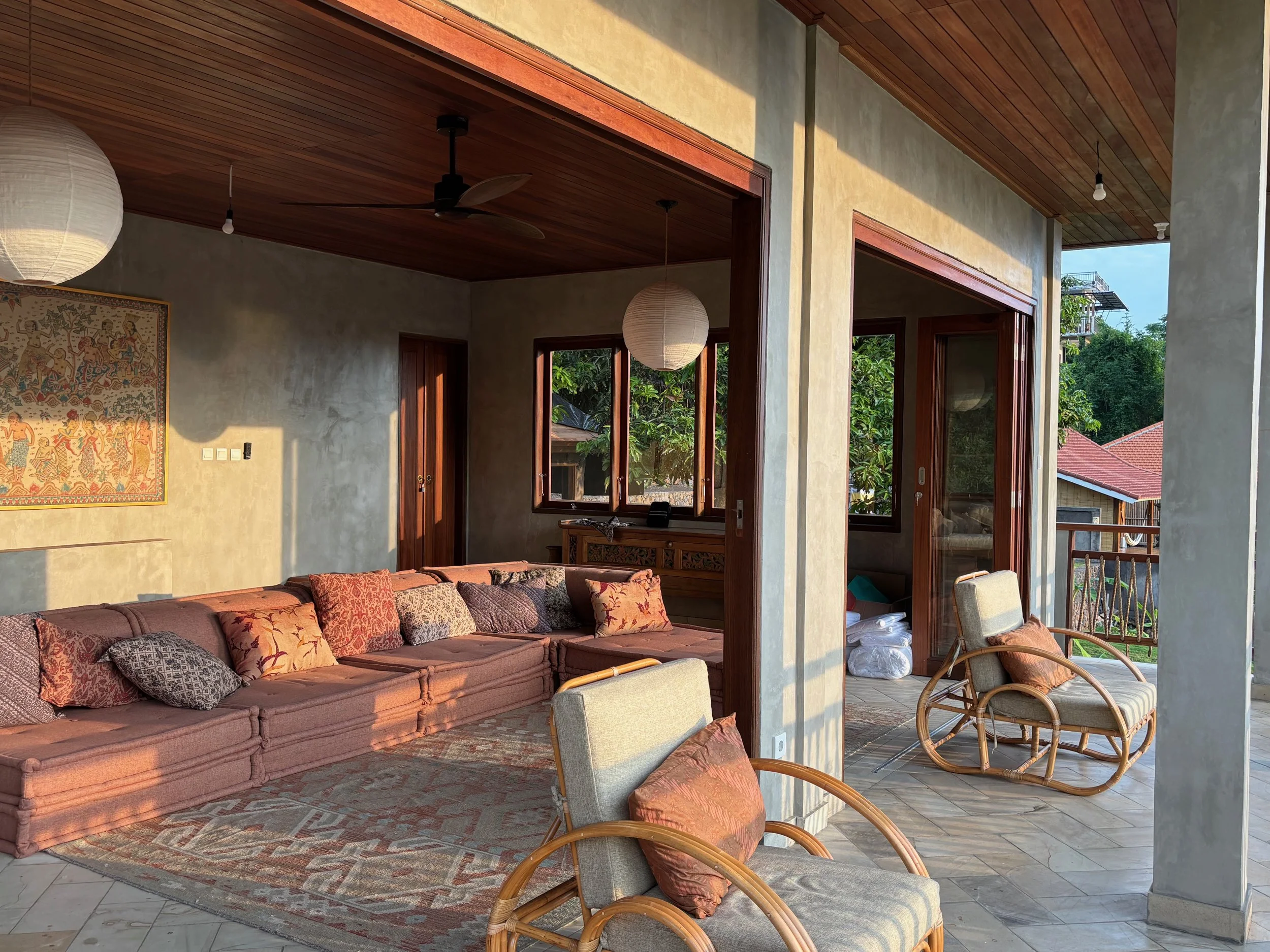 Living room with a pink sectional sofa and multiple decorative cushions, a vintage rug, wooden ceiling, paper lanterns, and large windows and sliding doors showing greenery outside.