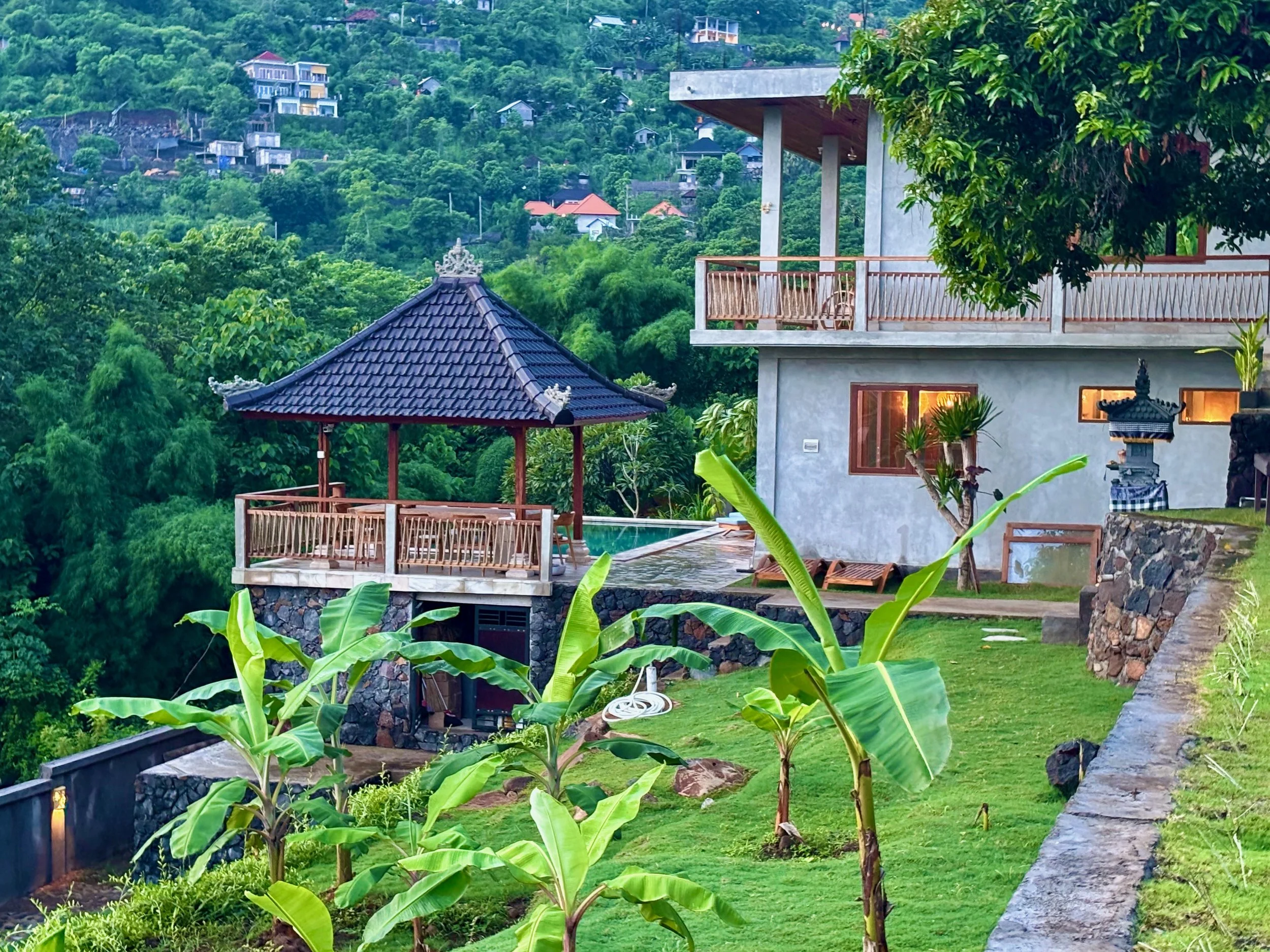 A hillside view of a modern house with a balcony, a traditional gazebo, a small garden with banana plants, and lush green vegetation in the background.