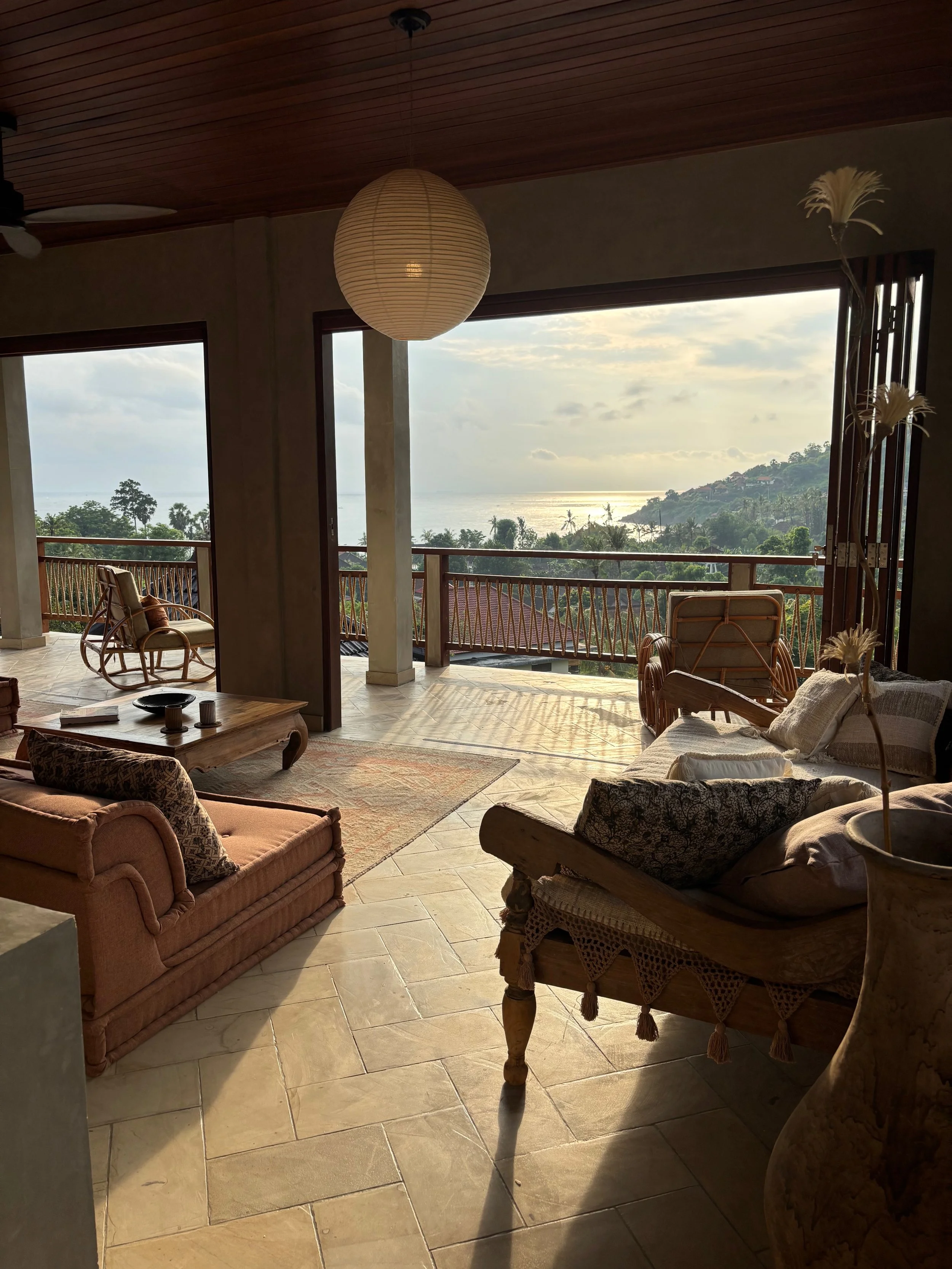Living room with large open balcony overlooking tropical landscape and ocean, furnished with vintage sofas, a wooden coffee table, rattan chairs, a hanging paper lantern, and decorative dried flowers.