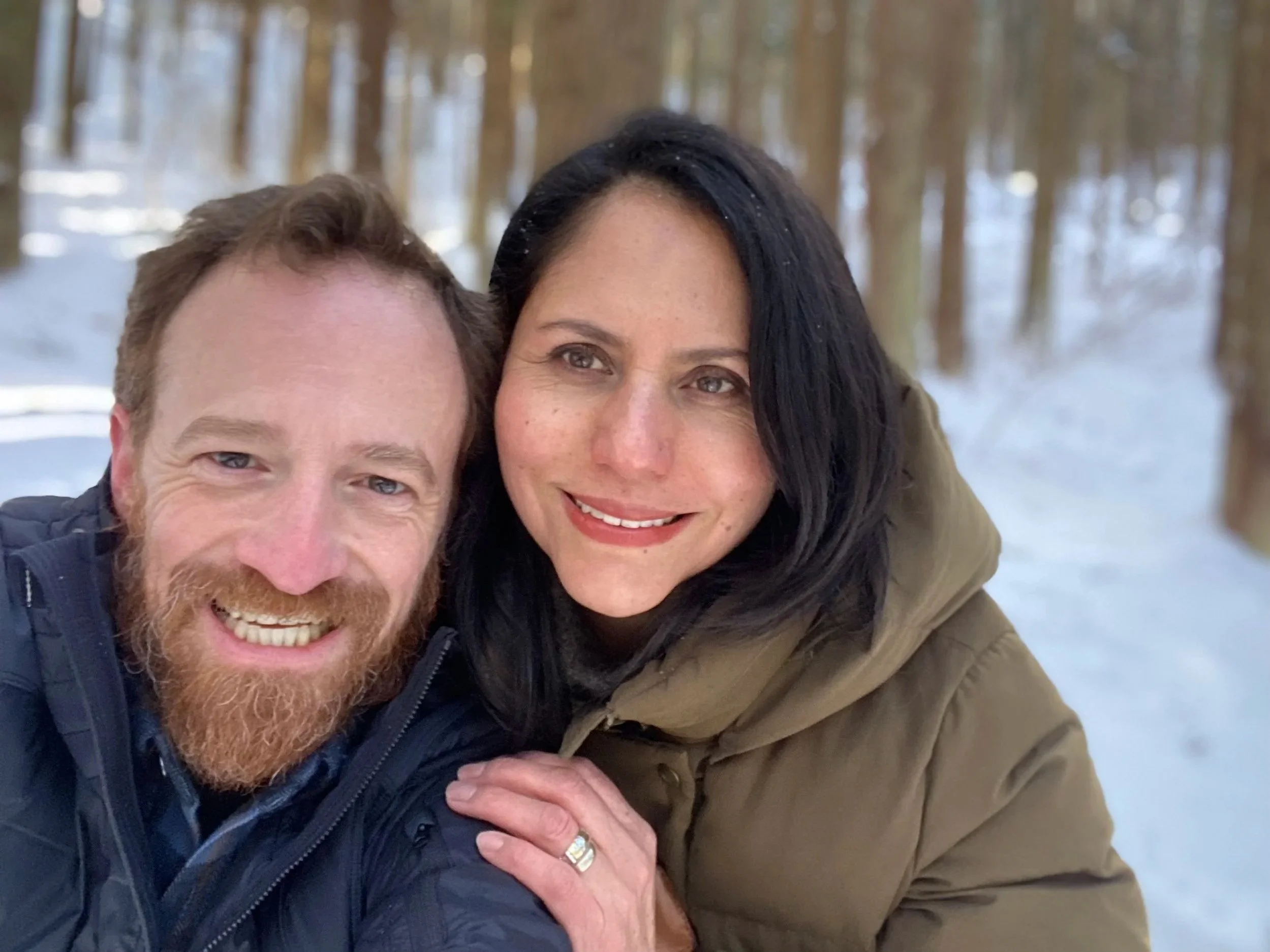 A smiling couple taking a selfie outdoors in a snowy forest with tall trees in the background.