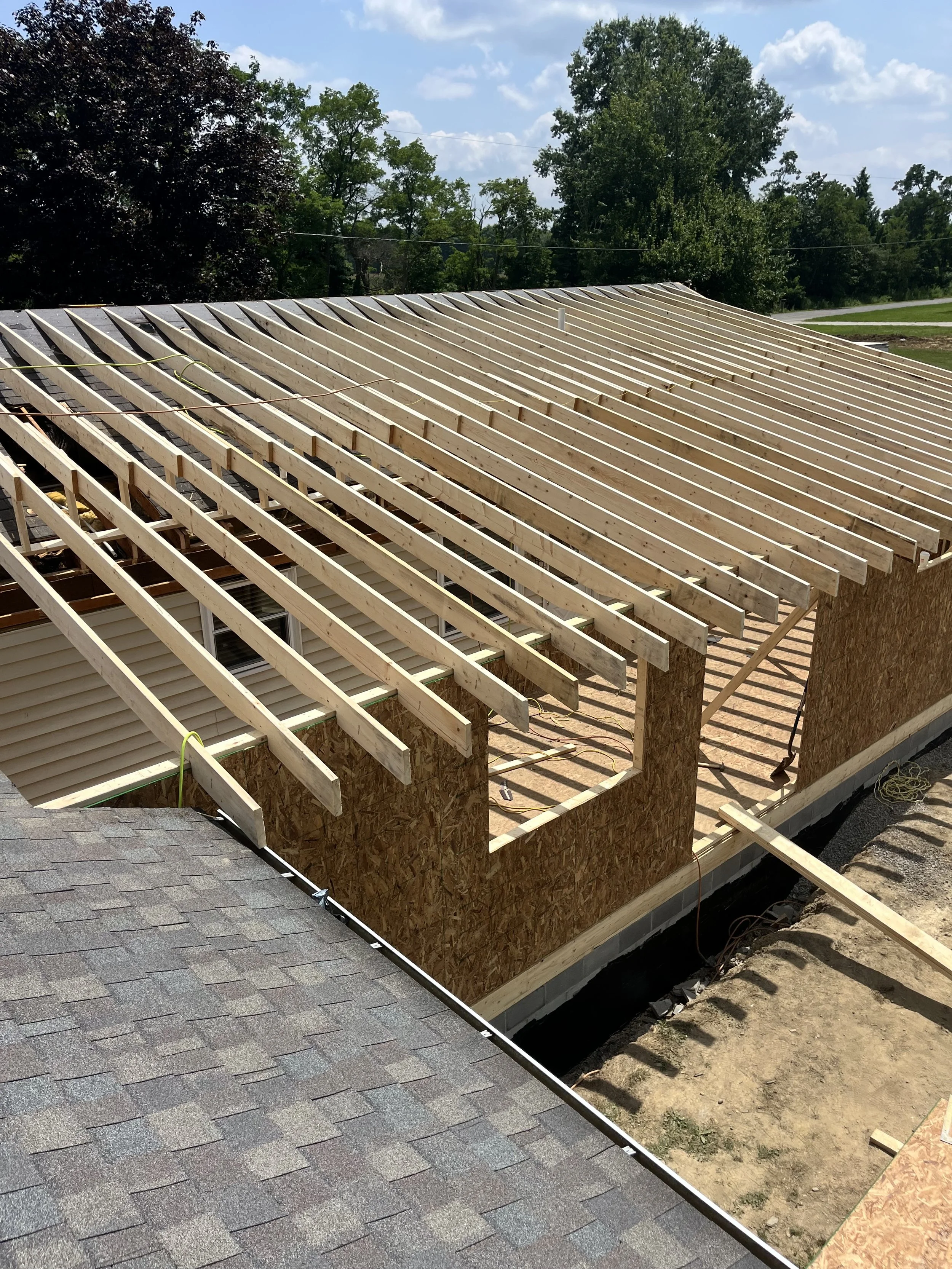 Construction site of a house with a roof under construction, showing wooden roof rafters and wall sheathing, with some neighboring rooftops and trees in the background.