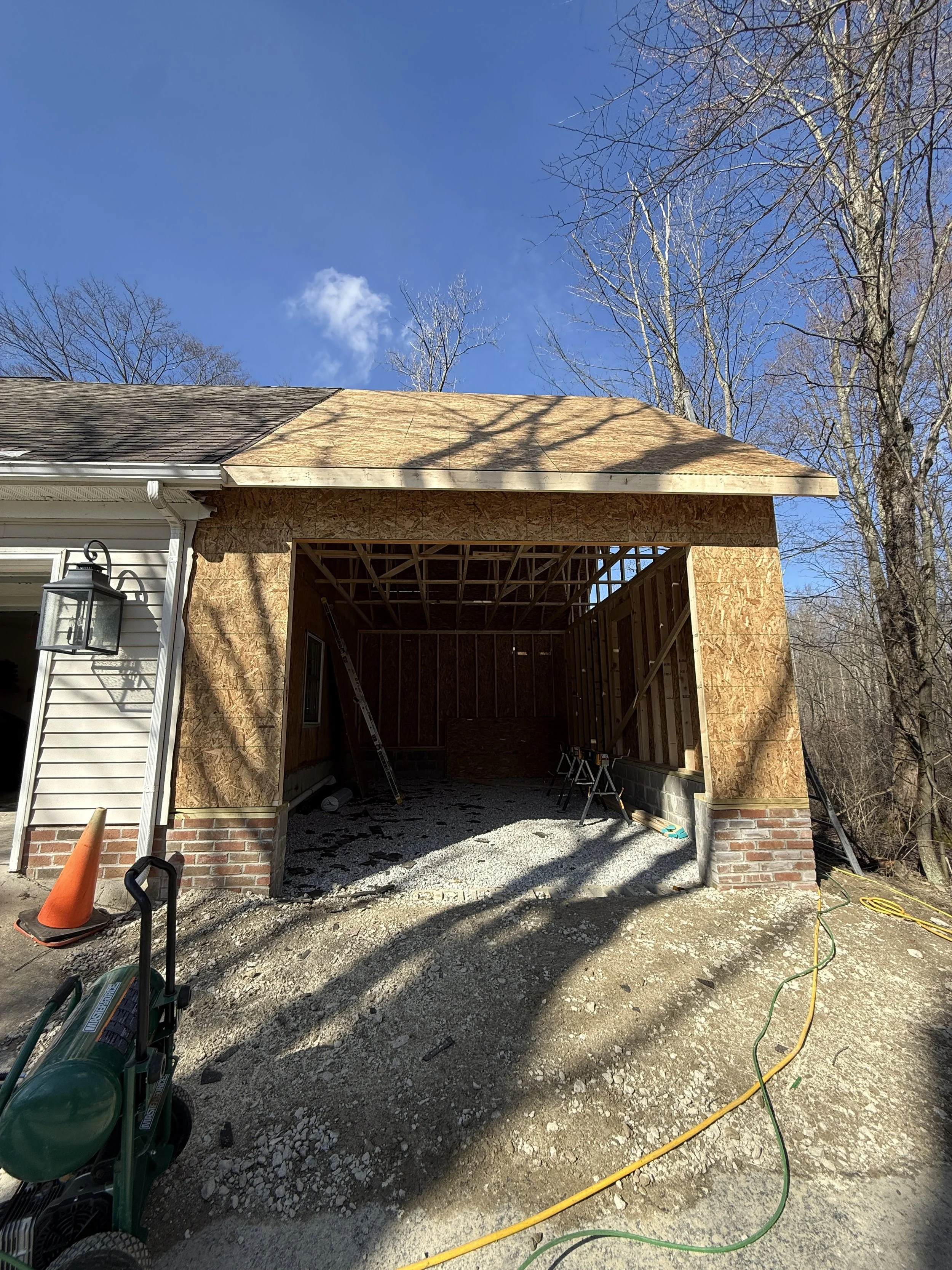 Construction of a garage addition to a house, with wooden framing and roofing in progress, on a sunny day with clear blue sky and leafless trees in the background.