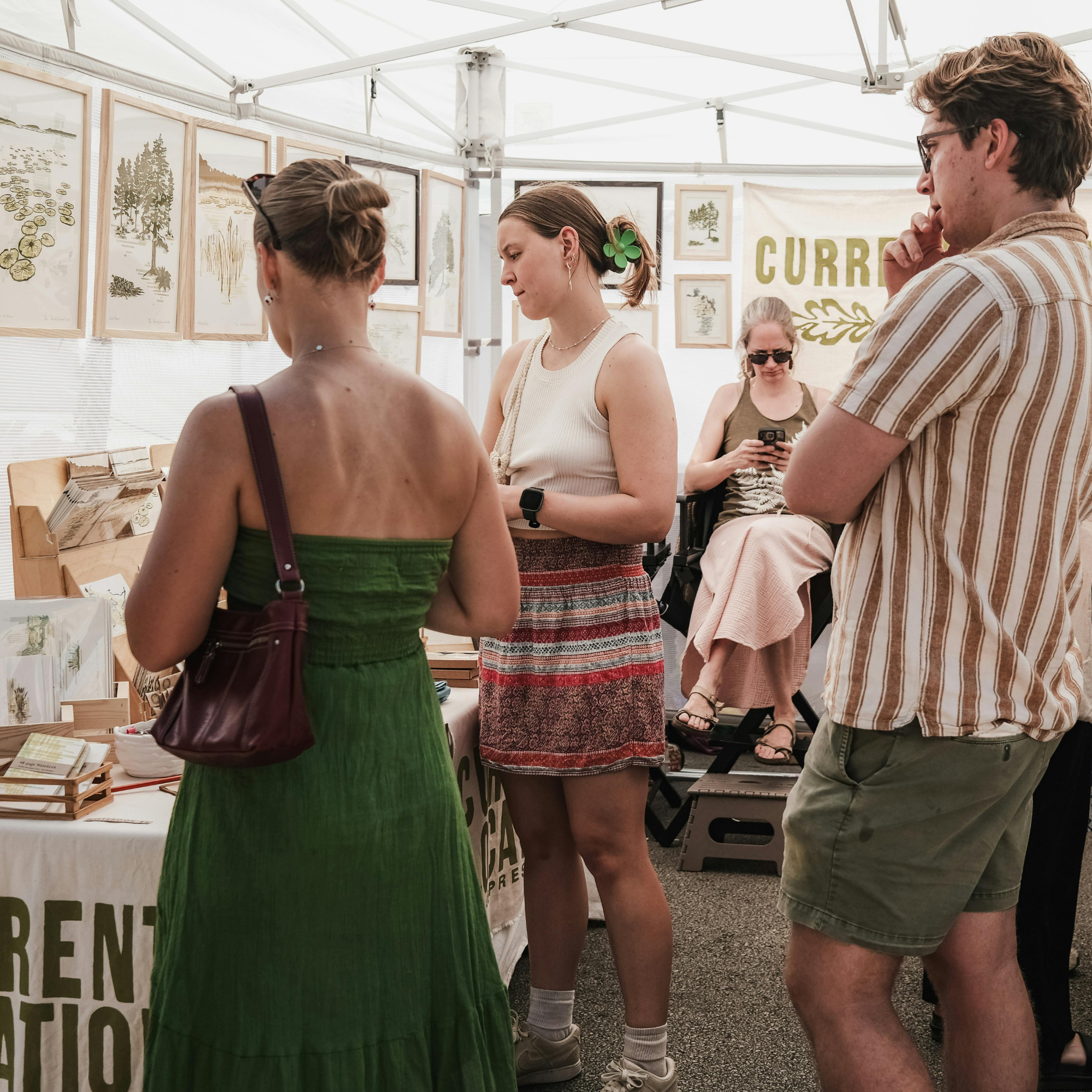 People browsing artwork at an outdoor art market and looking at framed pictures of trees and nature.