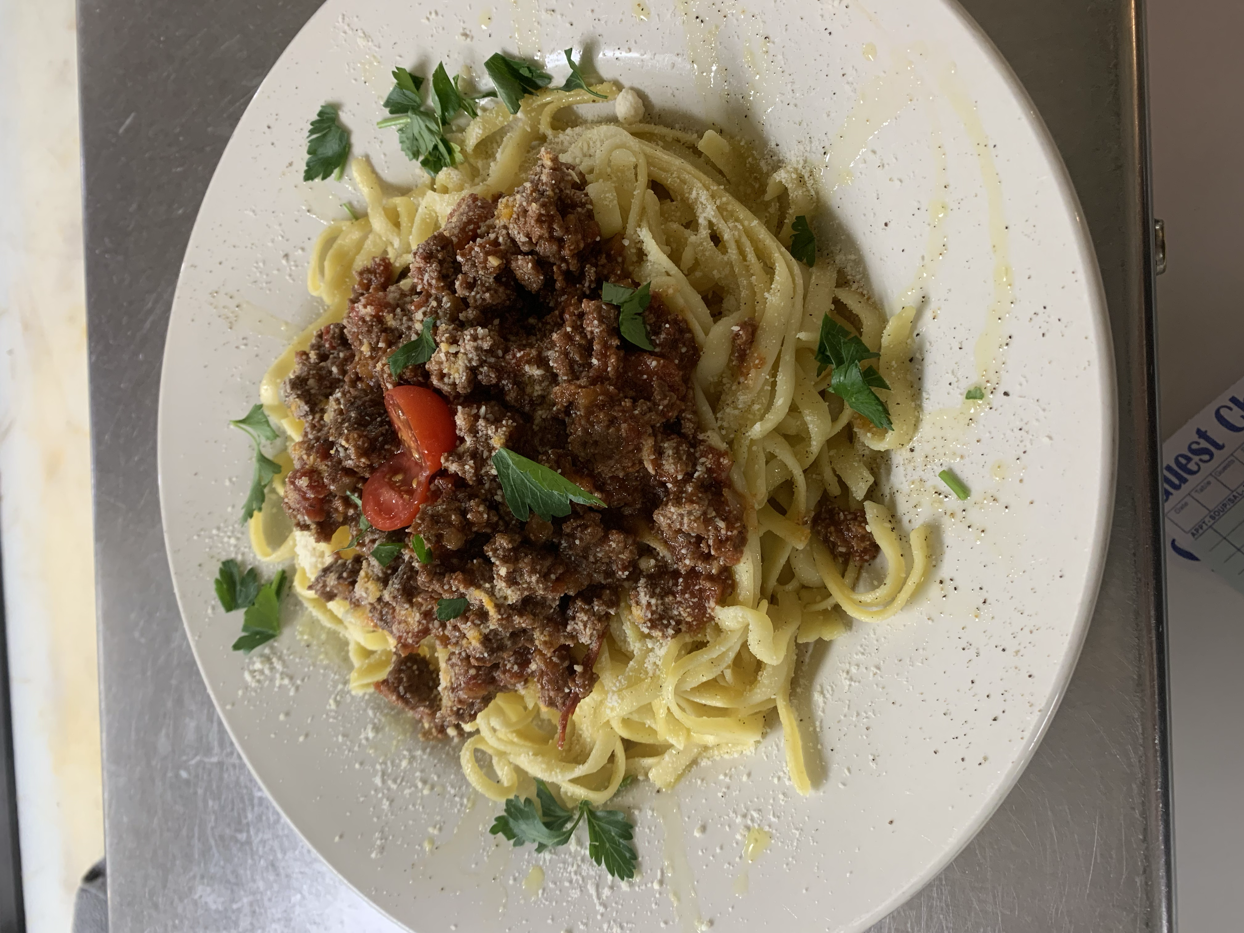 Plate of cooked spaghetti noodles topped with meat sauce, garnished with chopped parsley and slices of red pepper, with grated cheese and black pepper on the side.
