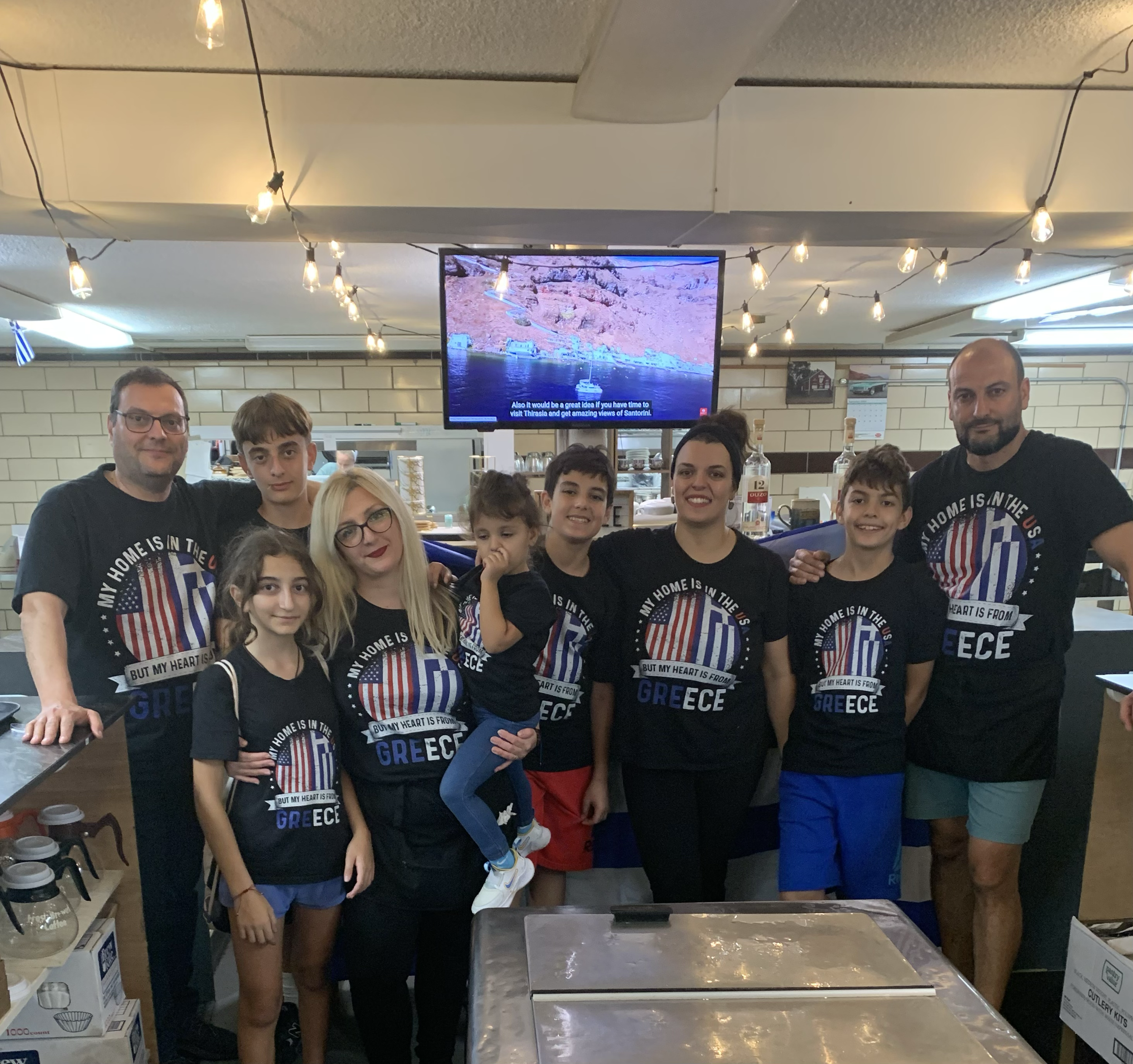 Family group photo in a restaurant, all wearing matching Greece-themed t-shirts with the American flag and Greek flag, with a TV screen in the background.