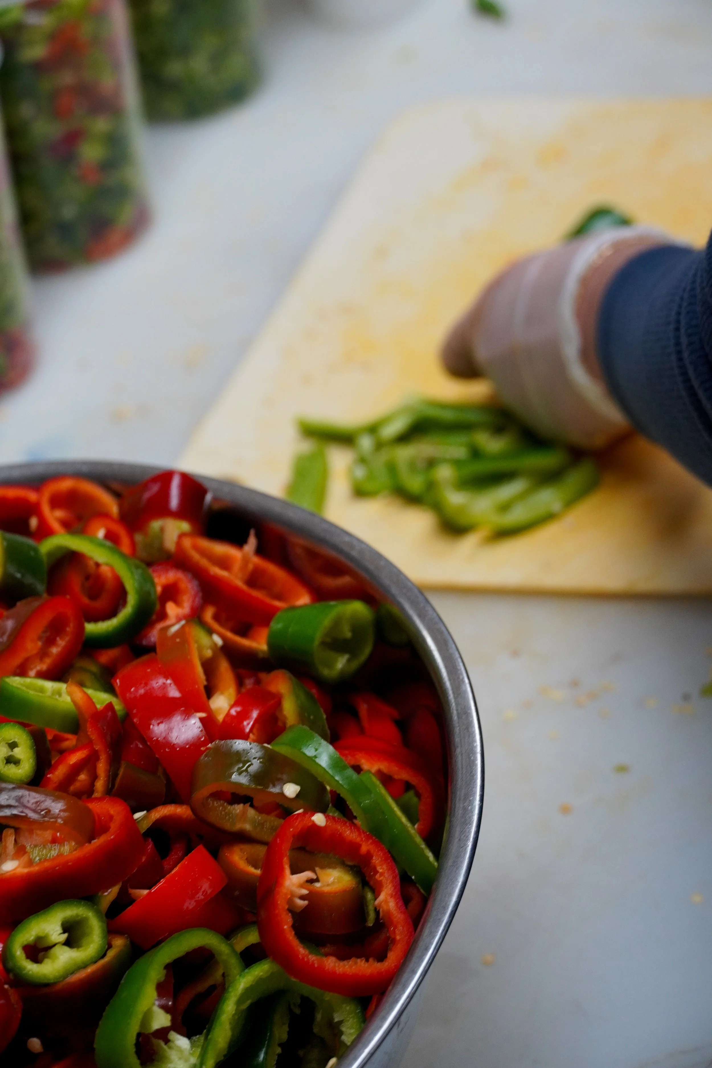 A bowl of sliced colorful red and green bell peppers on a white surface, with a hand chopping green bell peppers in the background.