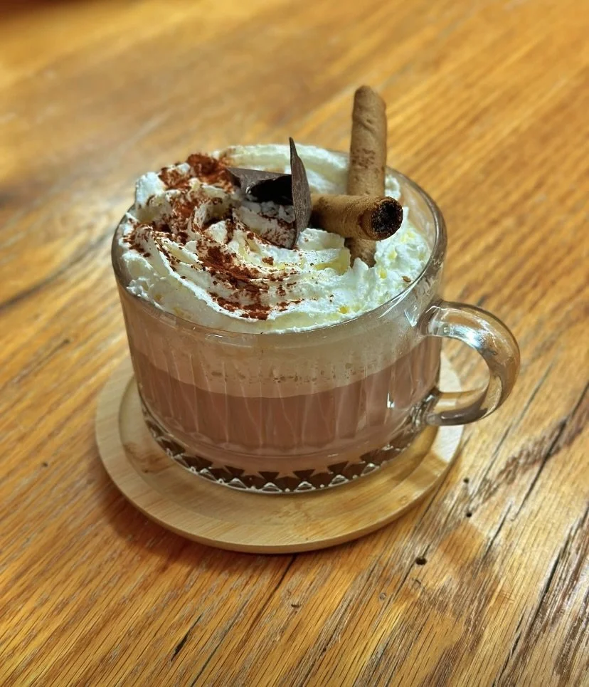 A glass cup of hot chocolate topped with whipped cream, chocolate shavings, and cinnamon sticks on a wooden table.