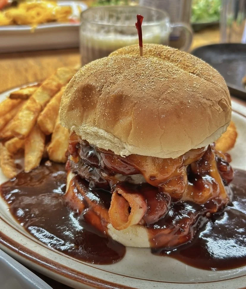 A meatloaf sandwich topped with barbecue sauce and a hamburger bun, served with French fries on a side. In the background, there is a beverage in a glass and some additional dishes.