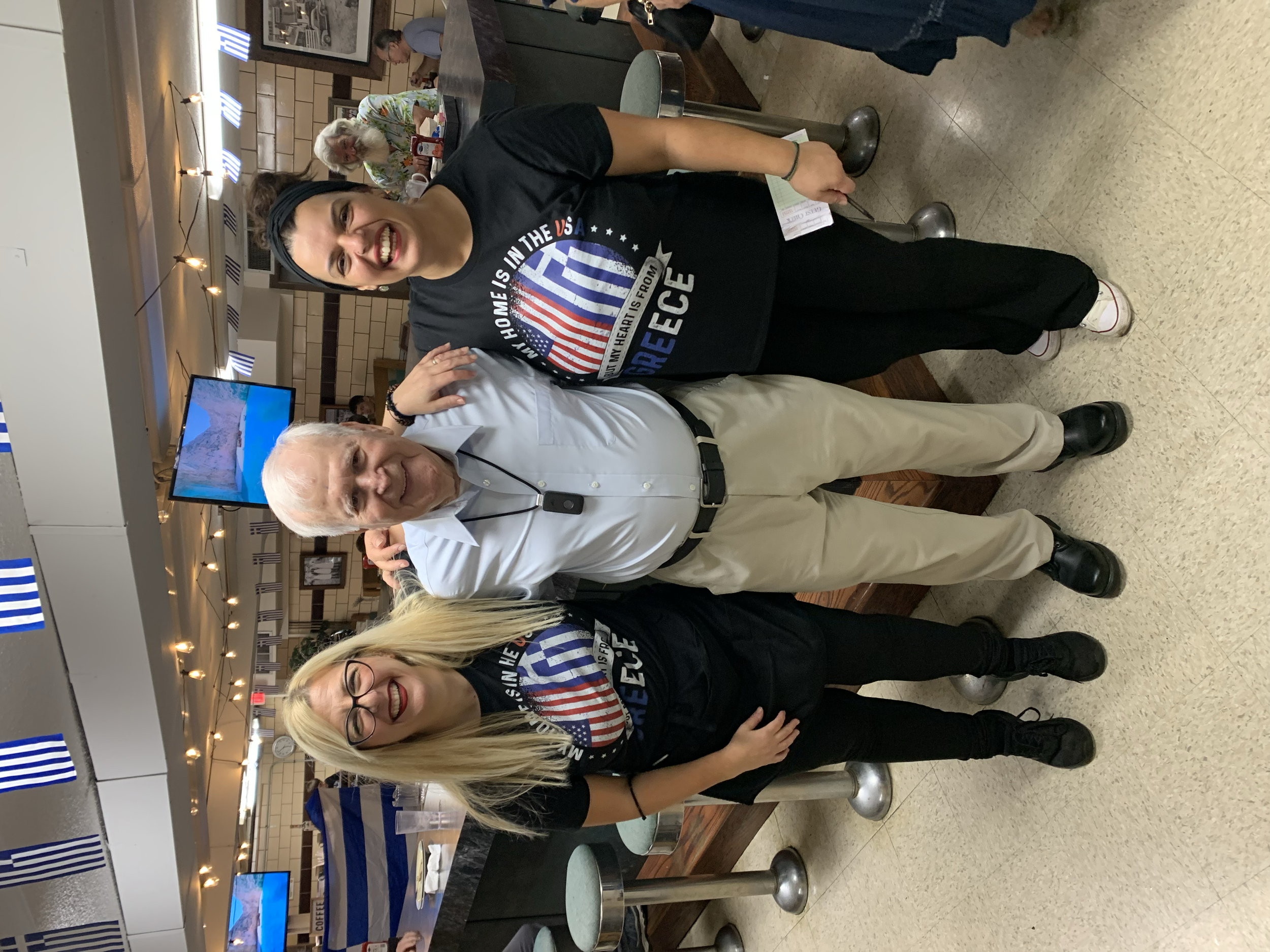 Three people standing together in a restaurant, with flags hanging from the ceiling. The woman in the front has blonde hair, glasses, and is wearing a black shirt with a patriotic design. The man in the middle has white hair, is wearing a white shirt