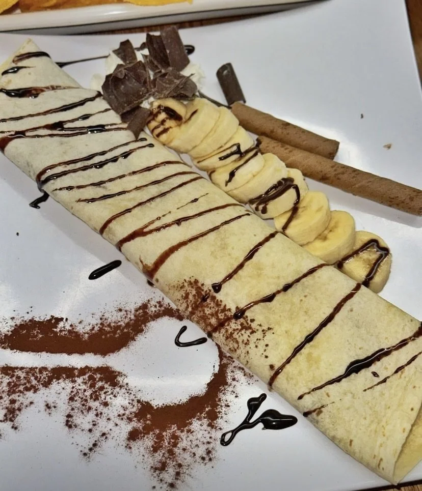 Dessert crepe filled with bananas, topped with chocolate shavings, drizzled with chocolate, served with cinnamon sticks and cocoa powder on the side.