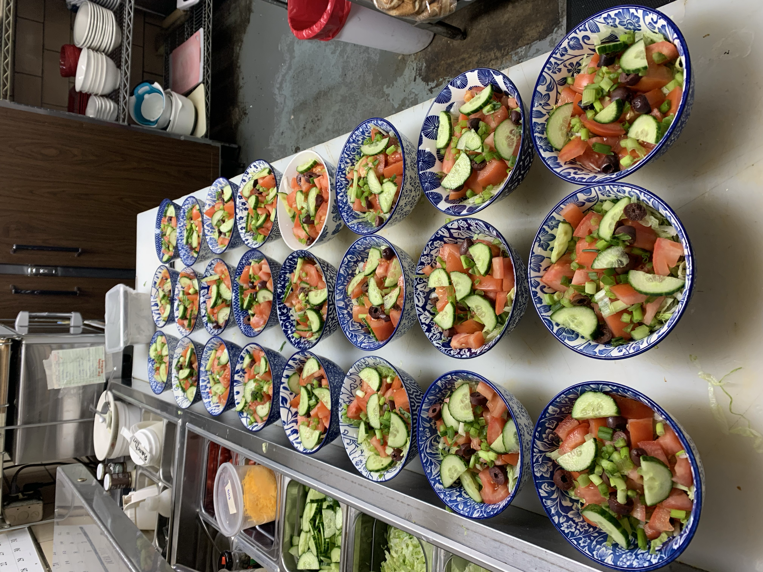 Multiple bowls of fresh vegetable salad arranged on a kitchen countertop, with chopped cucumbers, tomatoes, and olives visible.