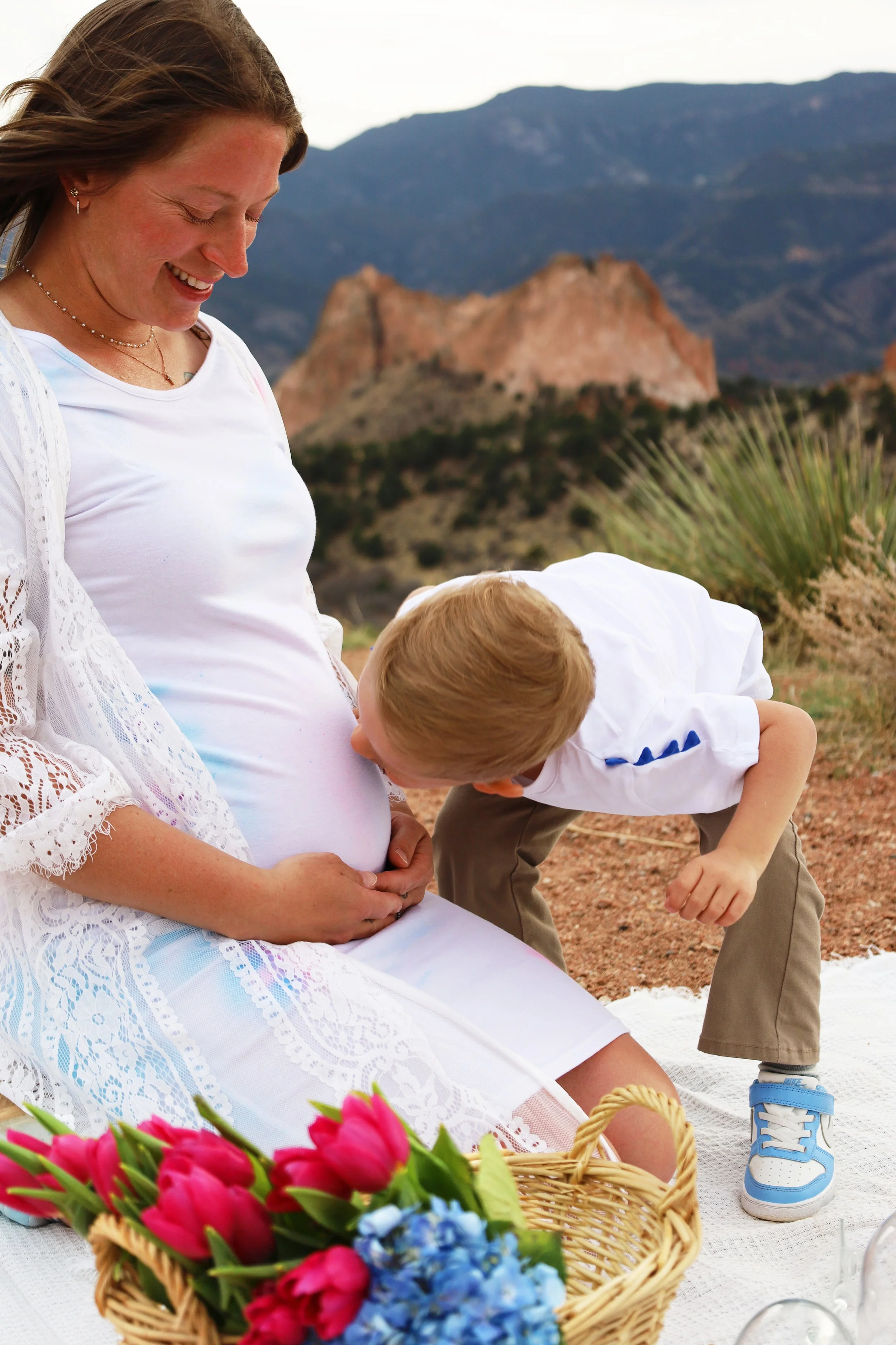 A woman with a pregnant belly sitting outdoors on a white blanket, smiling while a young boy kisses her belly, with a basket of pink and blue flowers in front, mountains in the background.