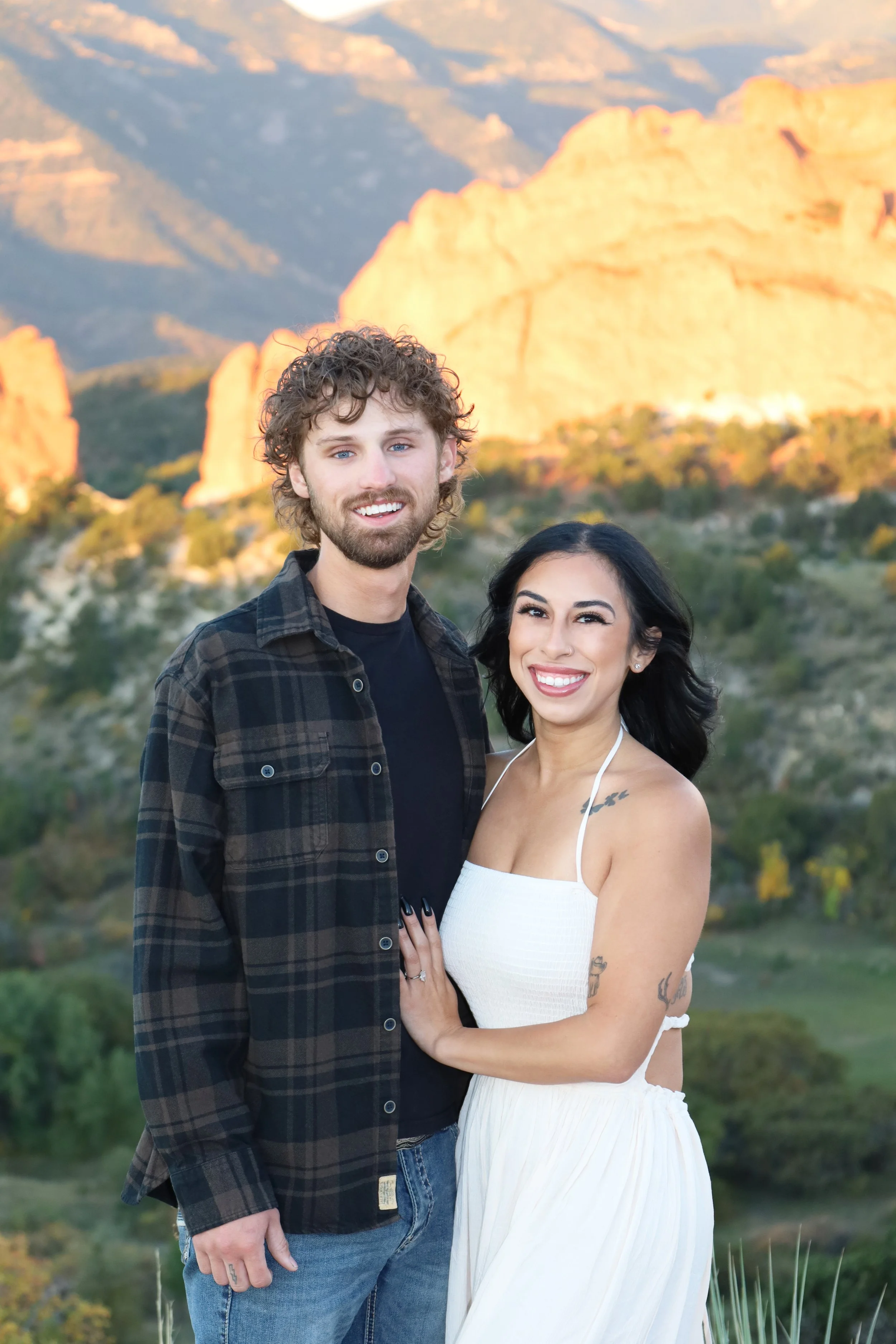 A smiling couple stands outdoors in front of a mountainous landscape with rocky formations at sunset.