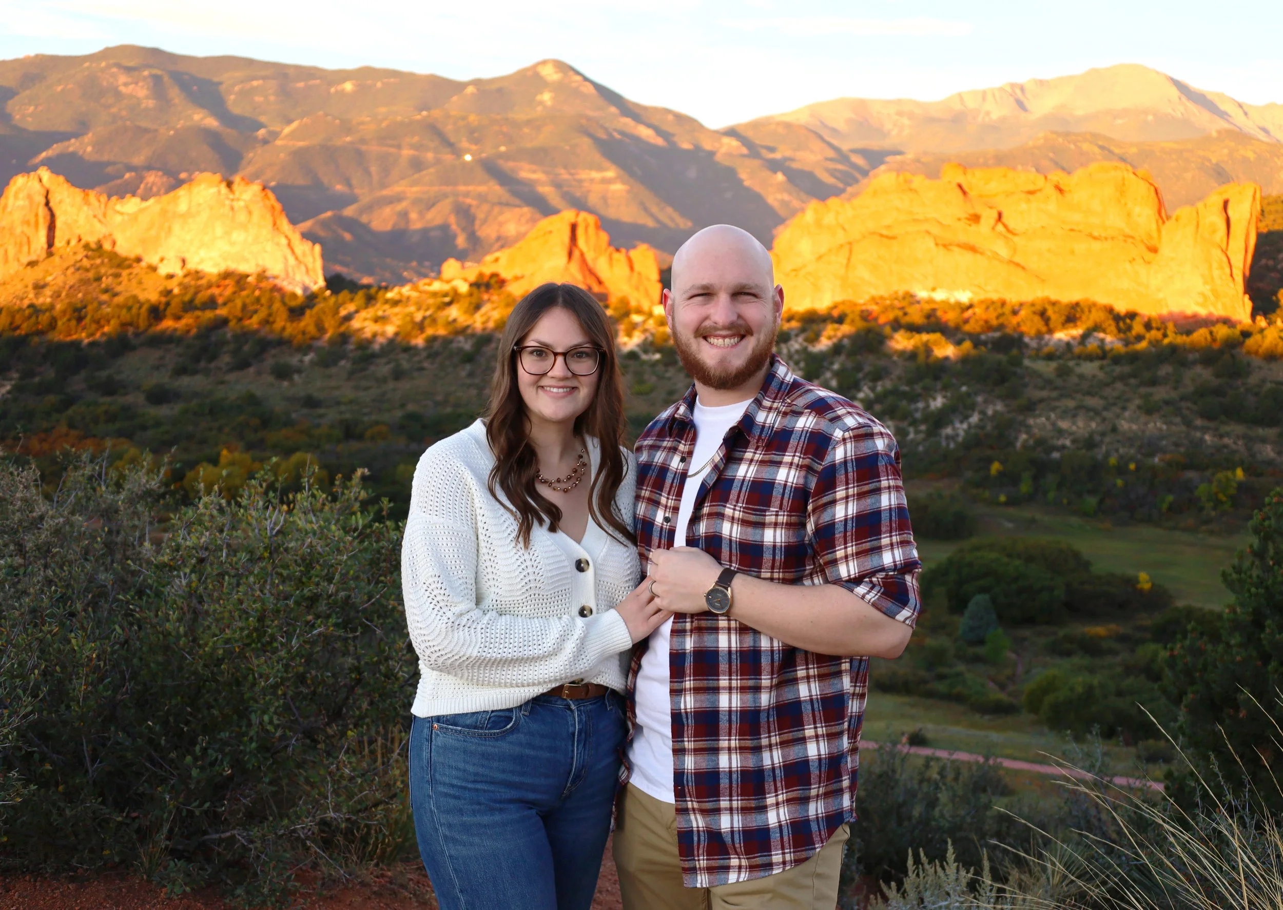 A smiling man and woman posing outdoors with mountains and red rock formations in the background during sunset.