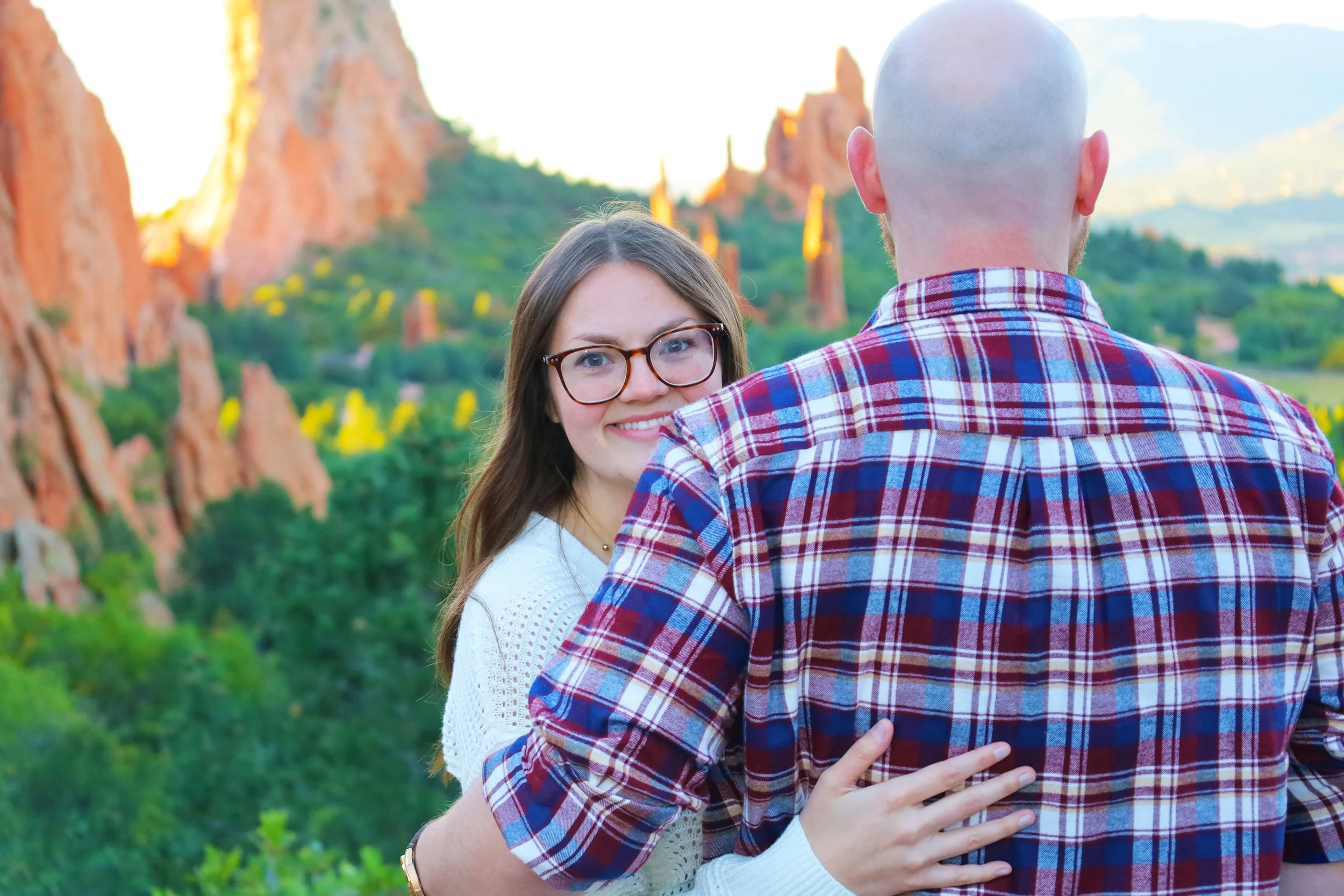 A couple embracing with their backs to the camera in front of red rock formations and green trees.
