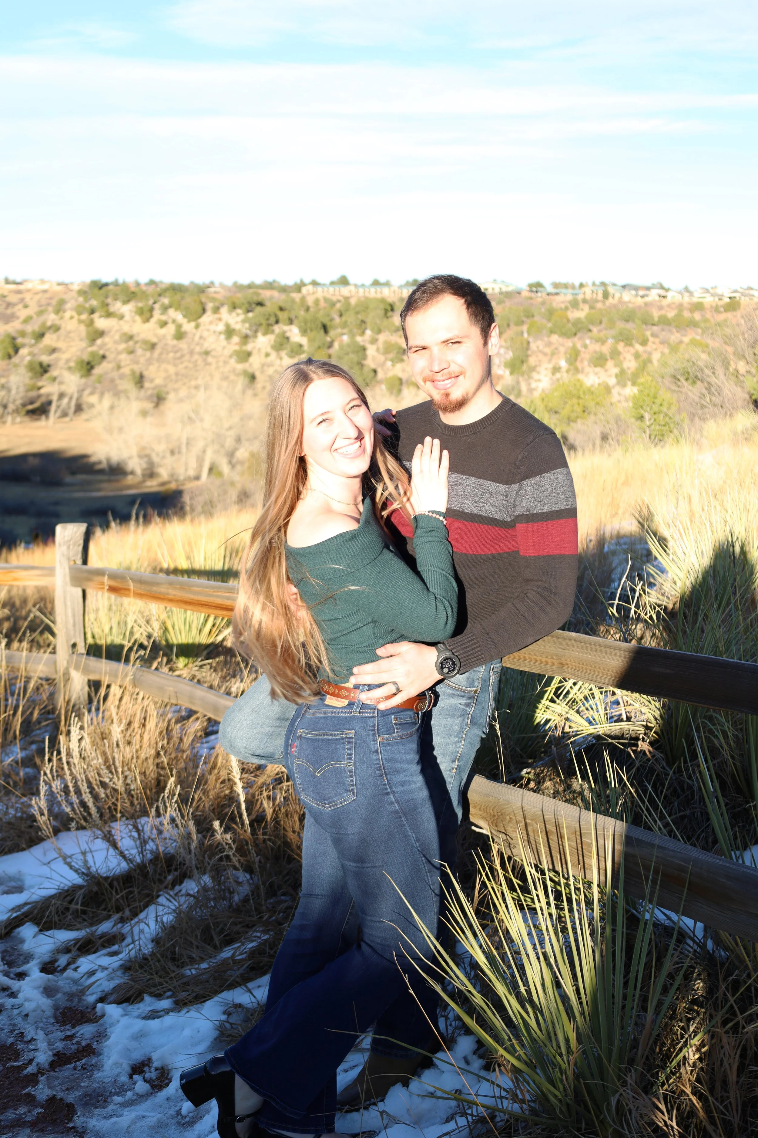 A smiling young couple embracing outdoors near a wooden fence on a sunny day with a grassy landscape and trees in the background.